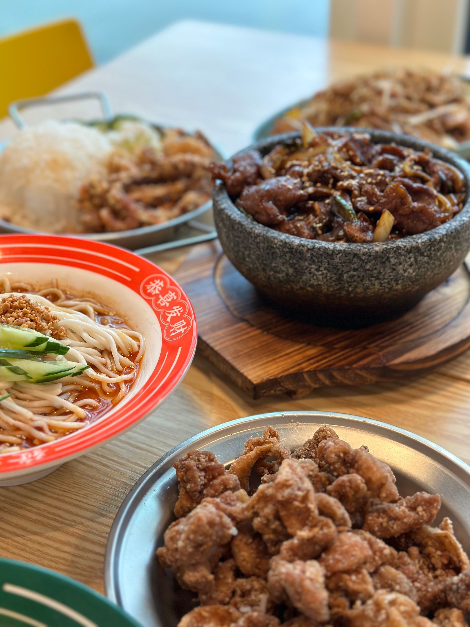 Assorted bowls of Asian noodle and meat dishes on a wooden table, including a red-rimmed soup bowl.