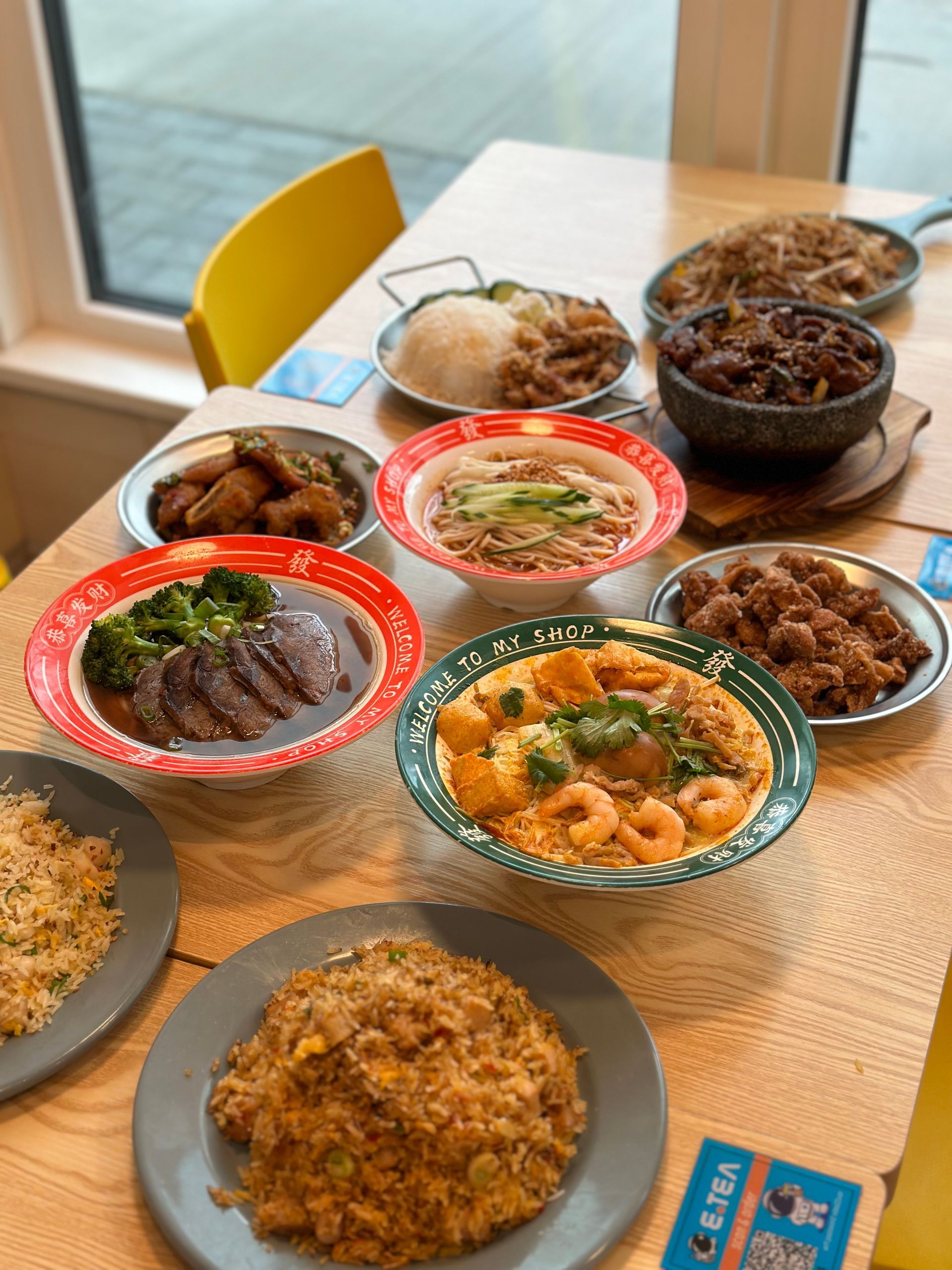 Assorted dishes on a wooden table, including rice, noodles, and meats in colorful bowls.
