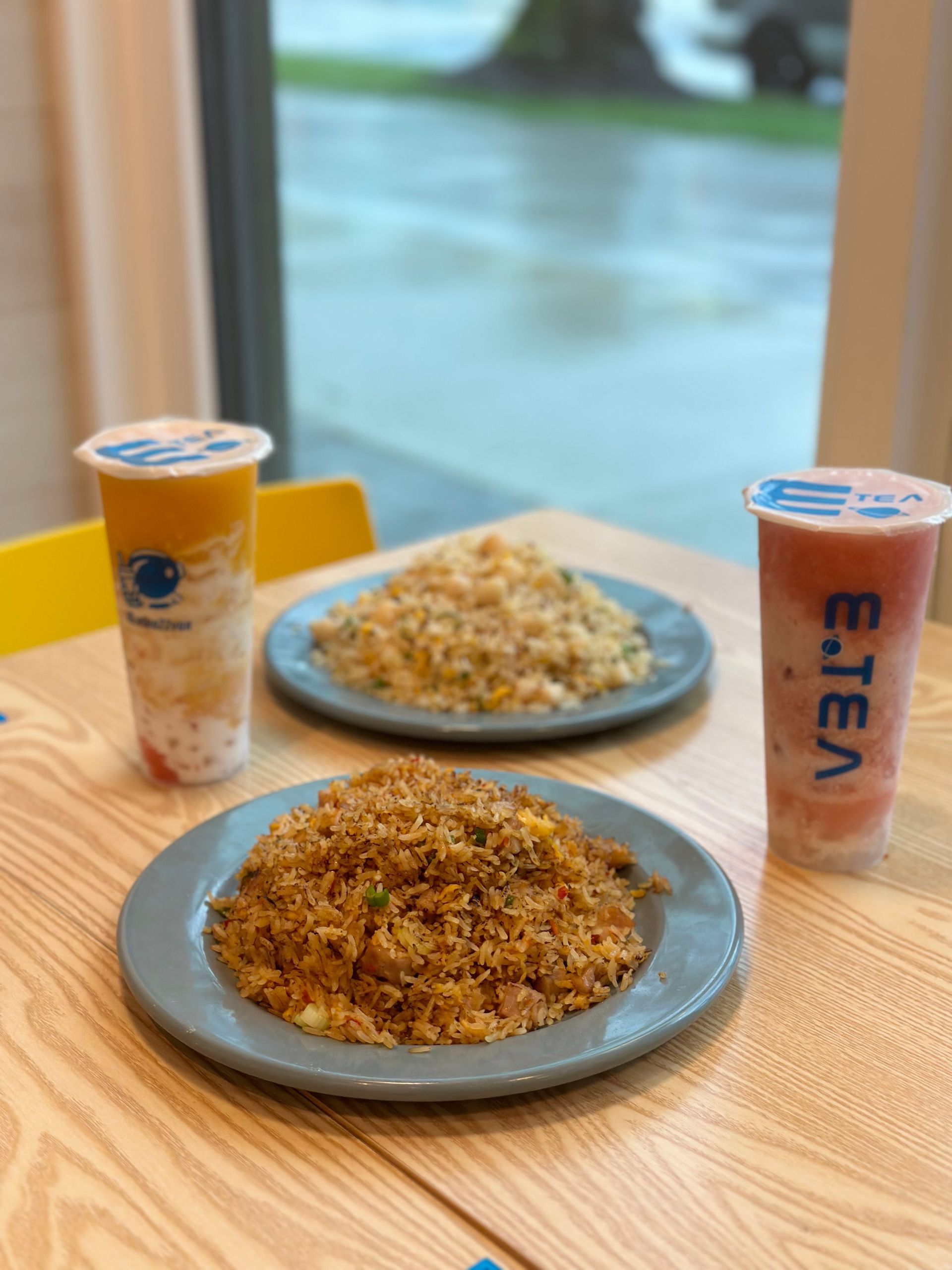 Two plates of rice and two drinks on a wooden table by a window