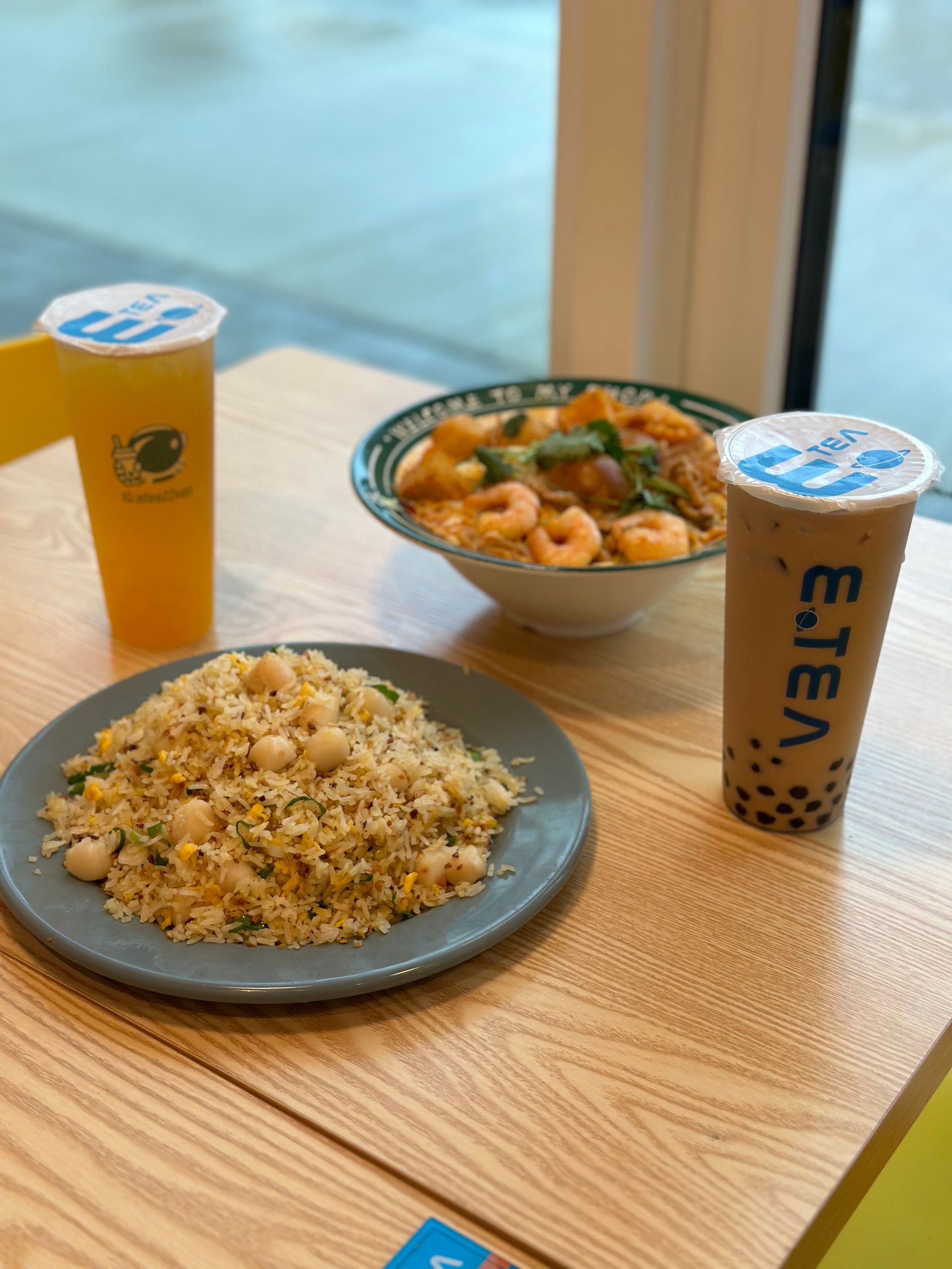 Plate of rice, bowl of curry, and two iced drinks on a wooden table by a window.