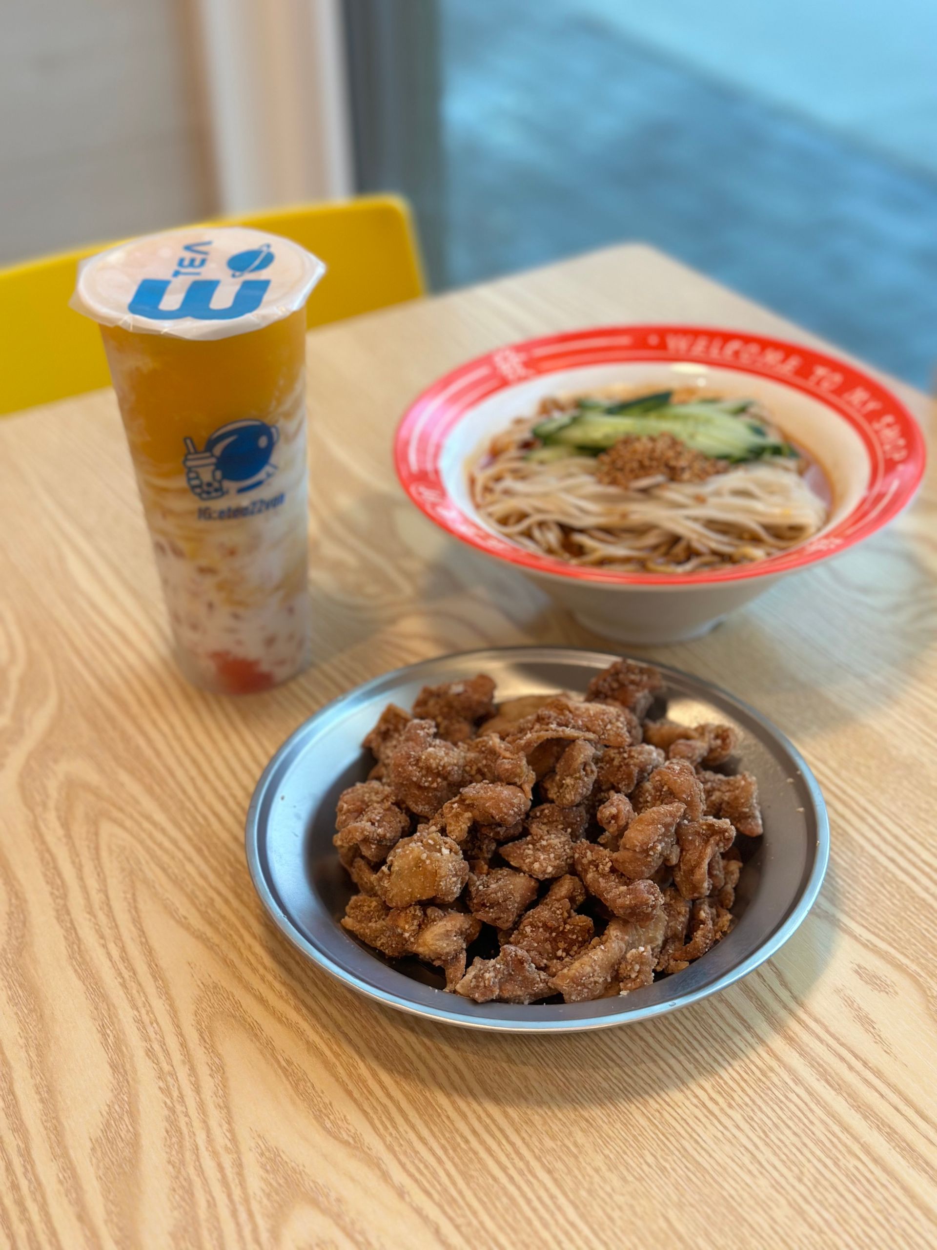 Tray with a bowl of noodles, fried chicken, and a yellow drink on a wooden table