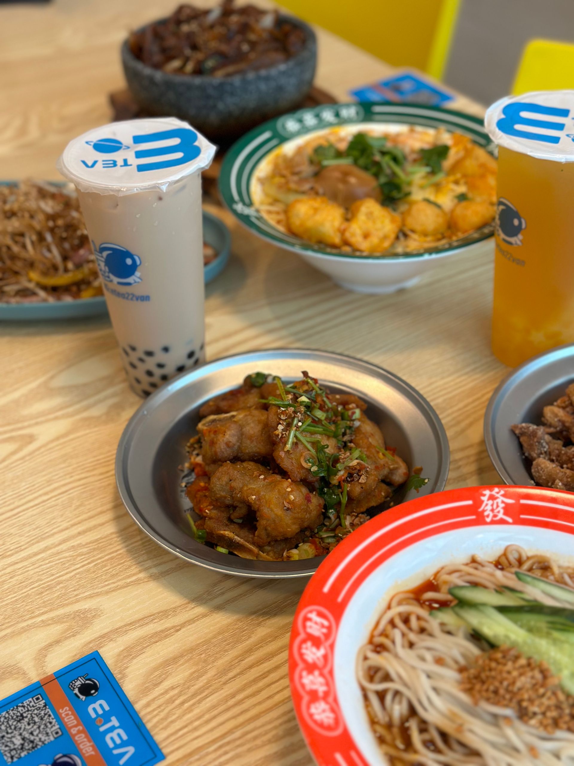 Table of Asian dishes and drinks on a wooden table, including noodle bowls and iced beverages