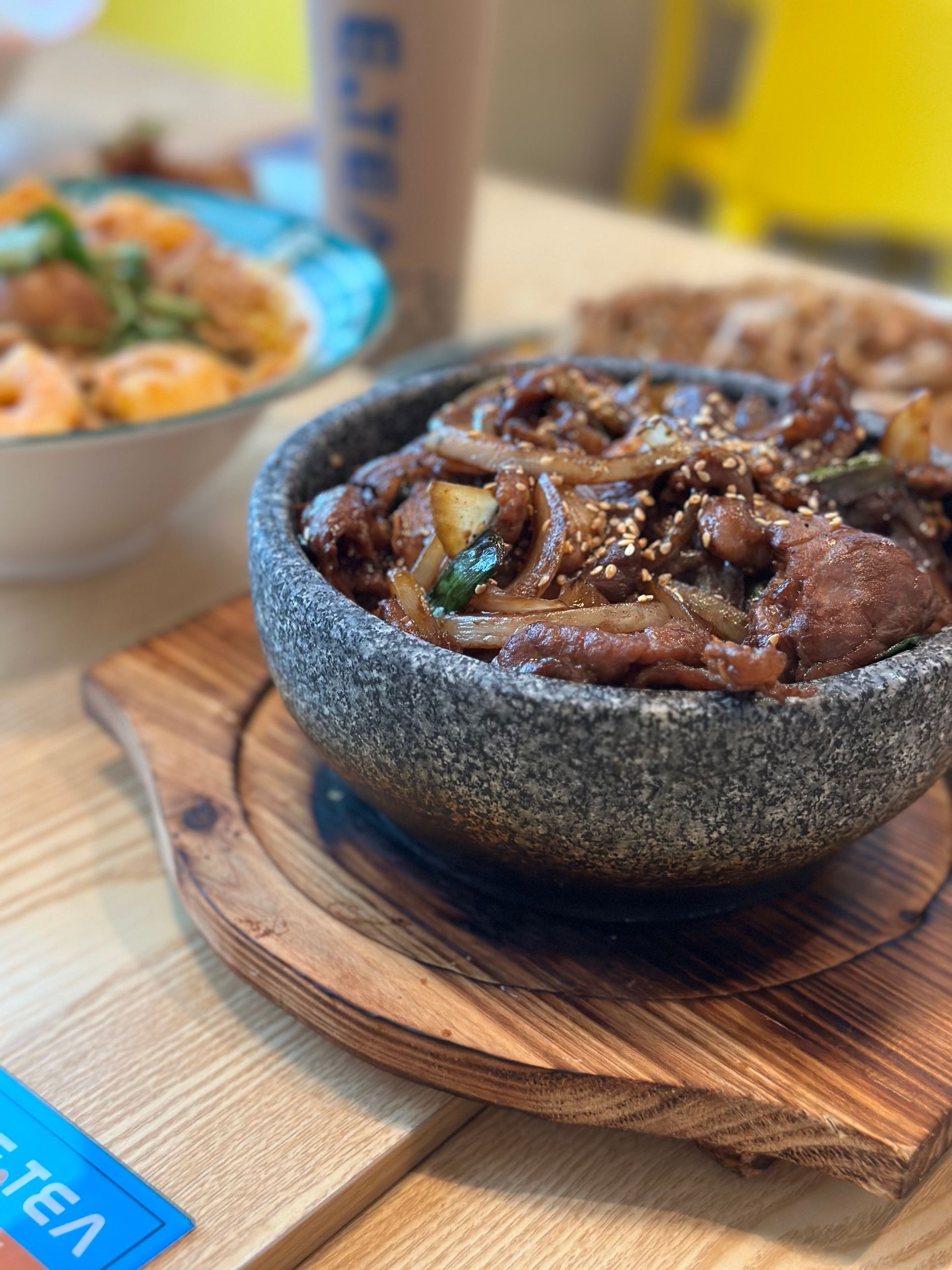 Blue bowl of dark sauced meat on a wooden board, with a blurred bowl of food in the background