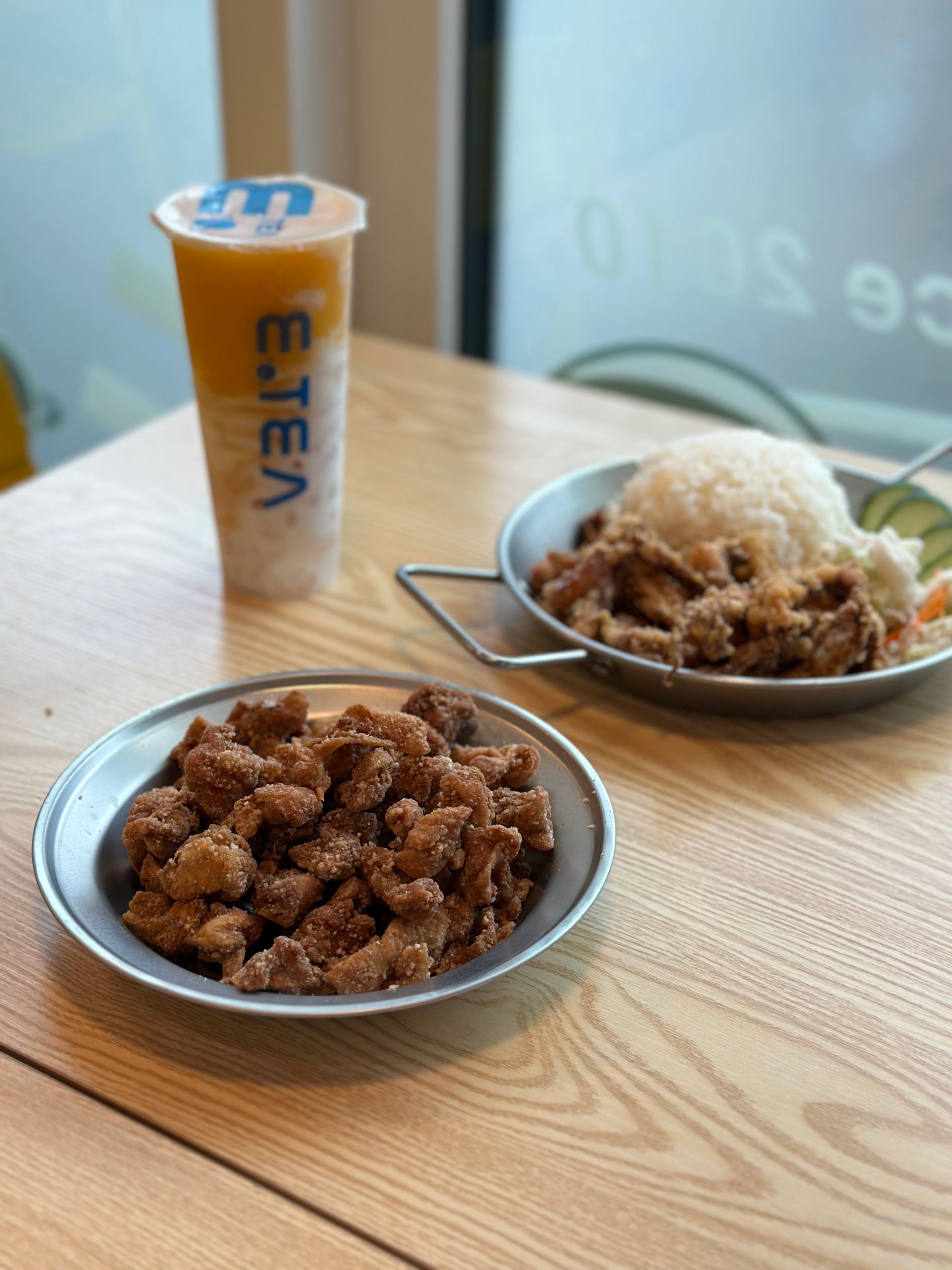 Two plates of fried chicken with a rice mound and a drink on a wooden table by a window