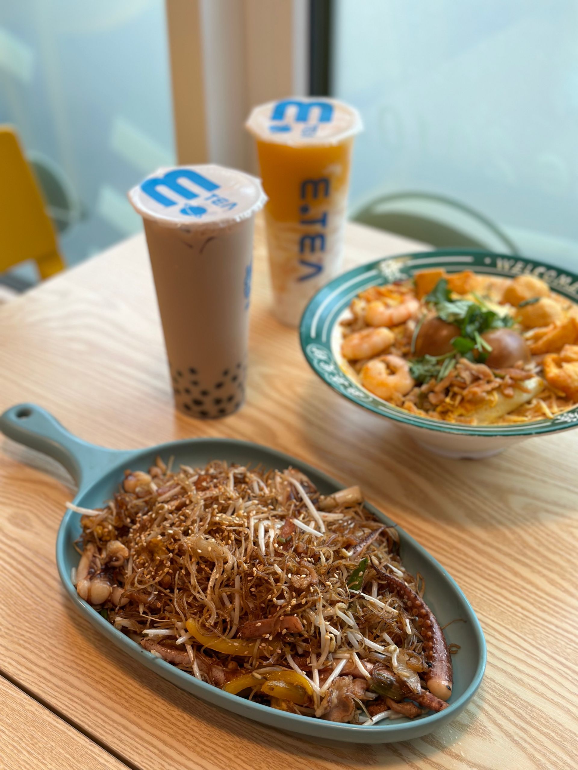 Two takeaway drinks and two noodle dishes on a wooden table by a window.