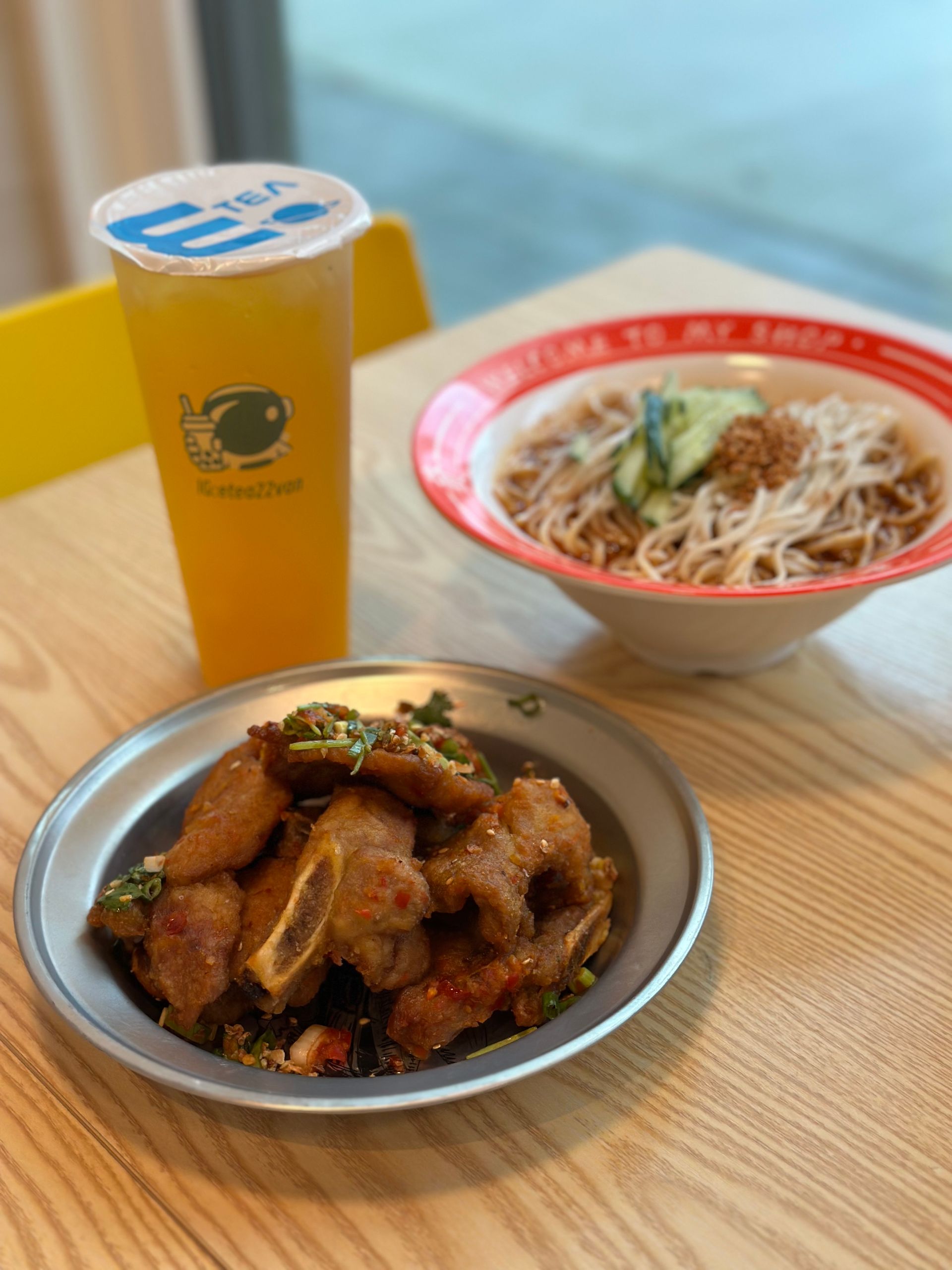 Meal tray with fried chicken, noodle soup, and a yellow drink on a table by a window