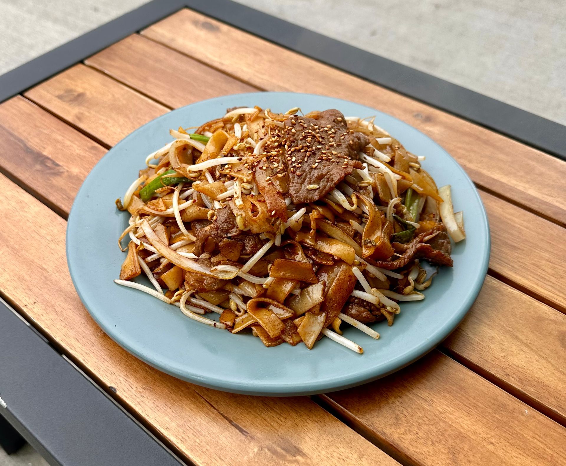 Plate of stir-fried noodles topped with sesame seeds and vegetables on a blue plate atop a wooden table