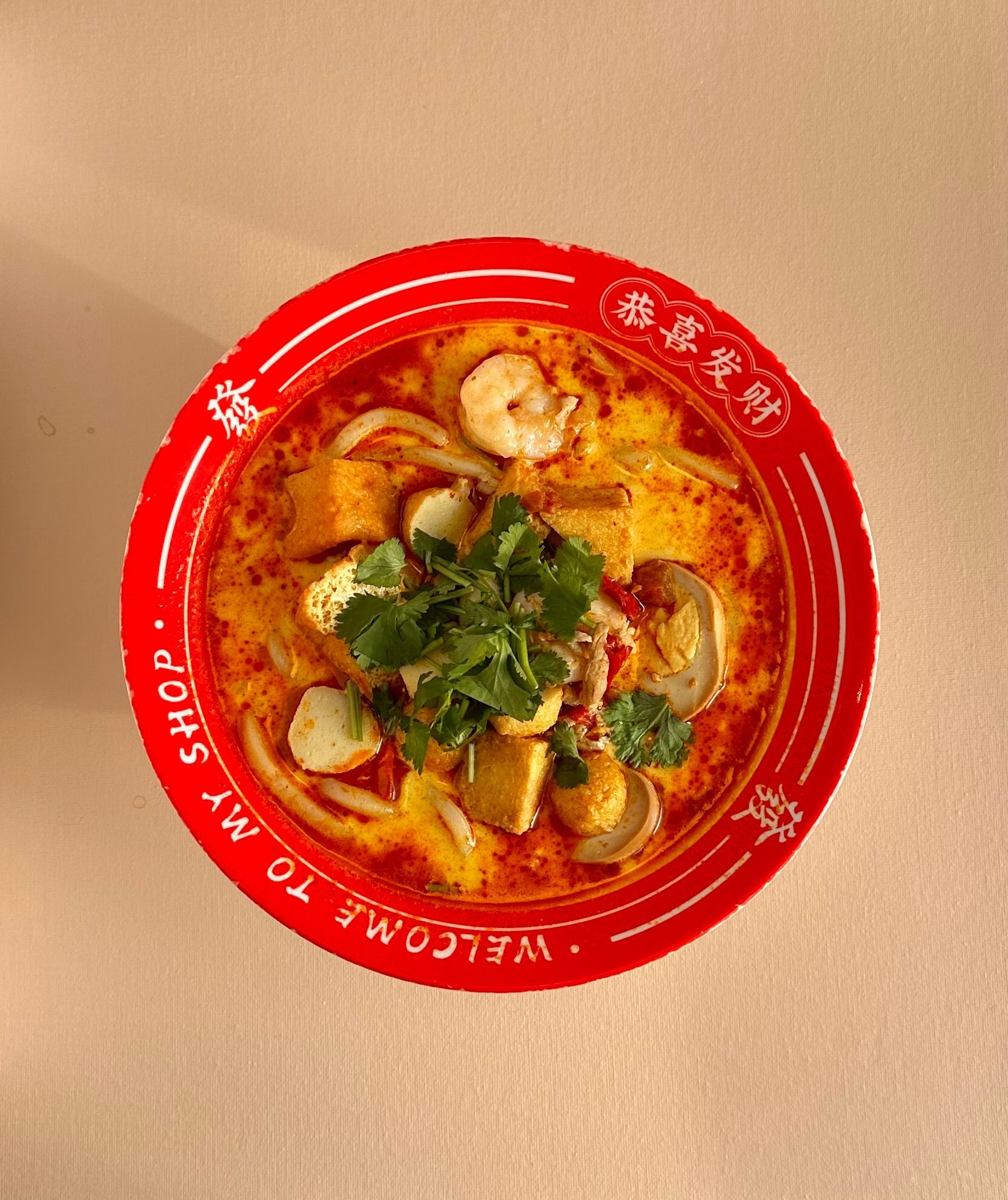 Red bowl of noodle soup with herbs and shrimp on a beige table, viewed from above