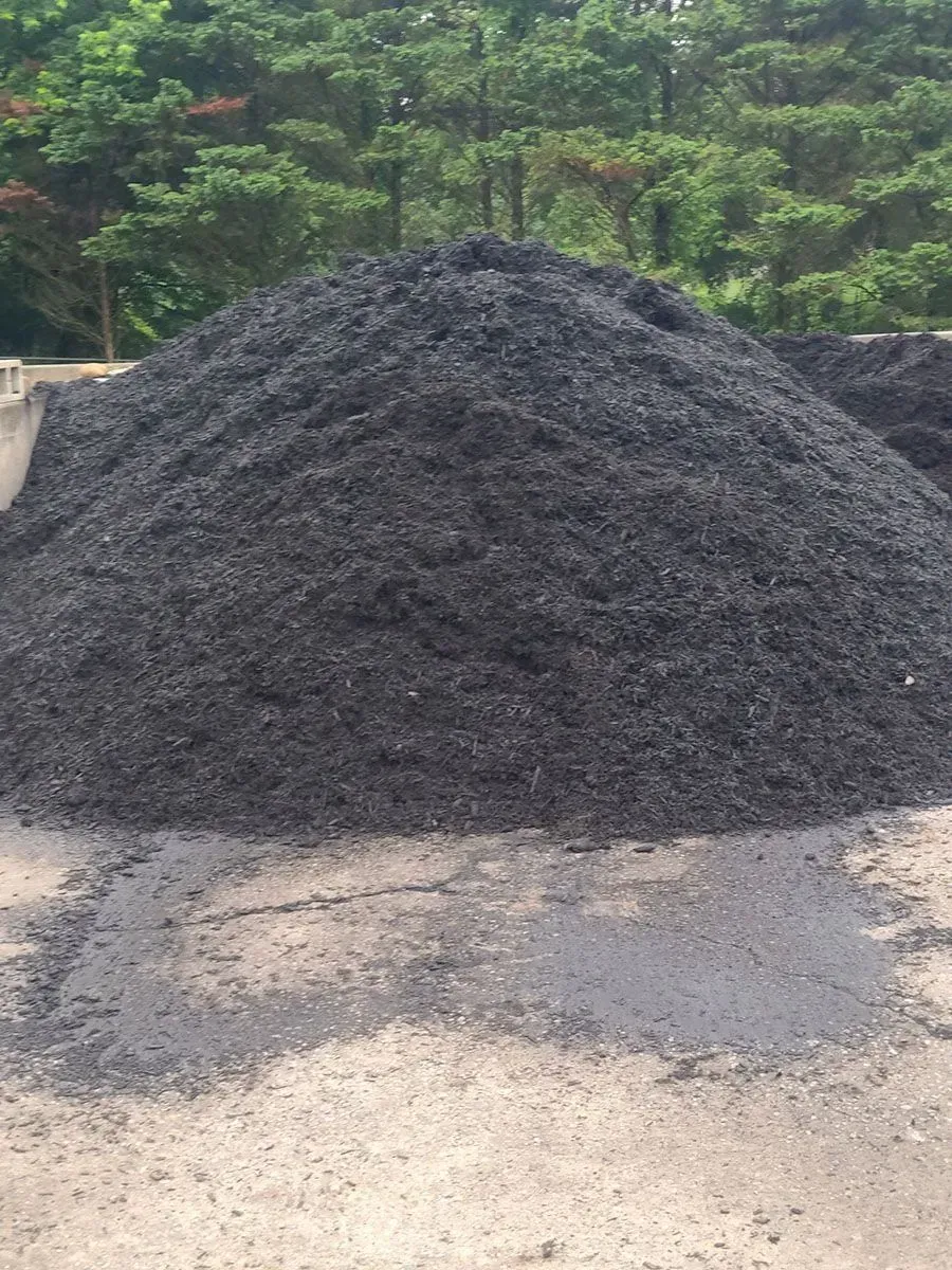 Large pile of dark gravel on a dirt lot with trees in the background