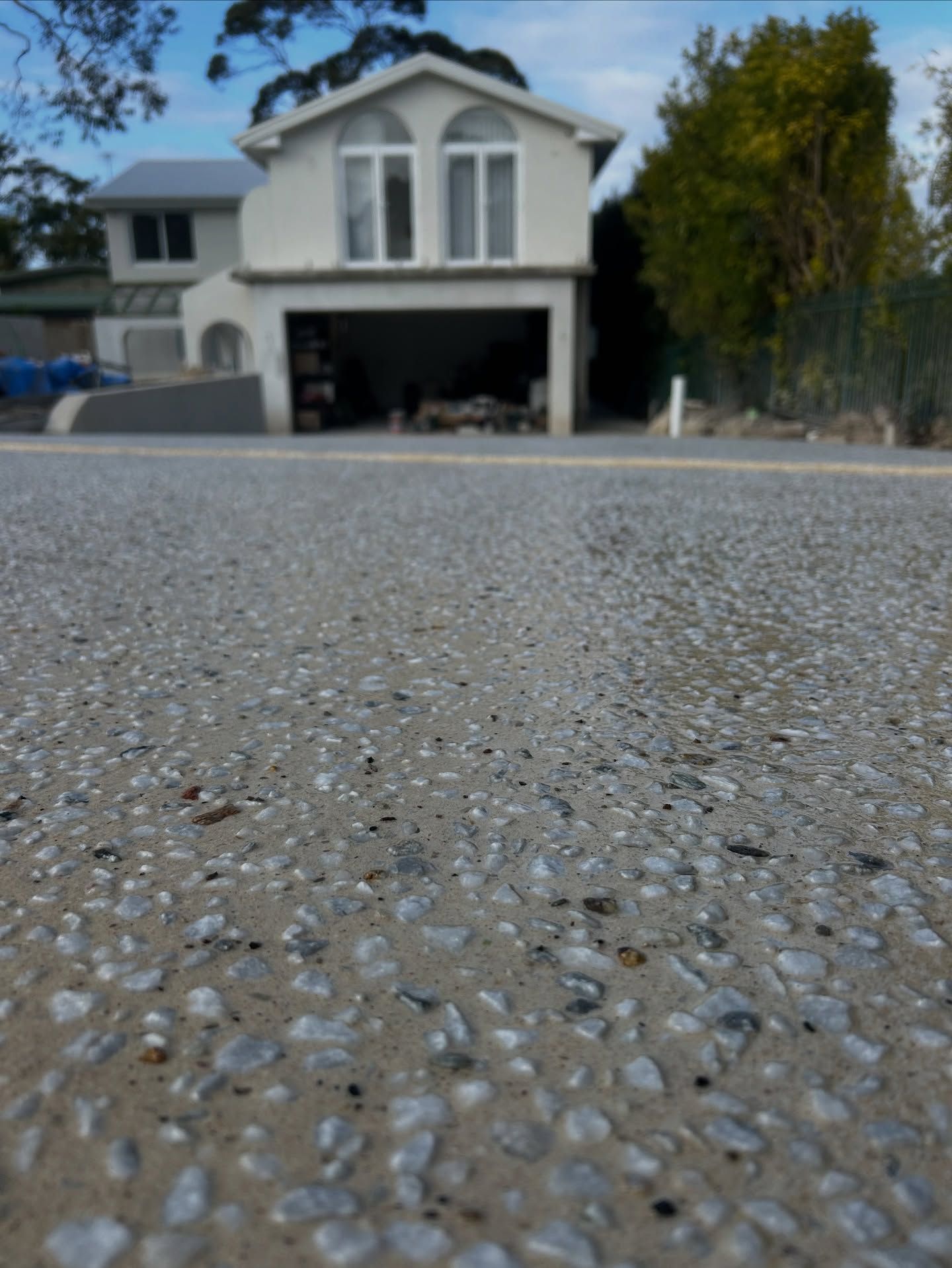 A low-angle view of a grey exposed aggregate driveway leading up to a two-story white house with an open garage. — JDB Concreting in Wollongong, NSW