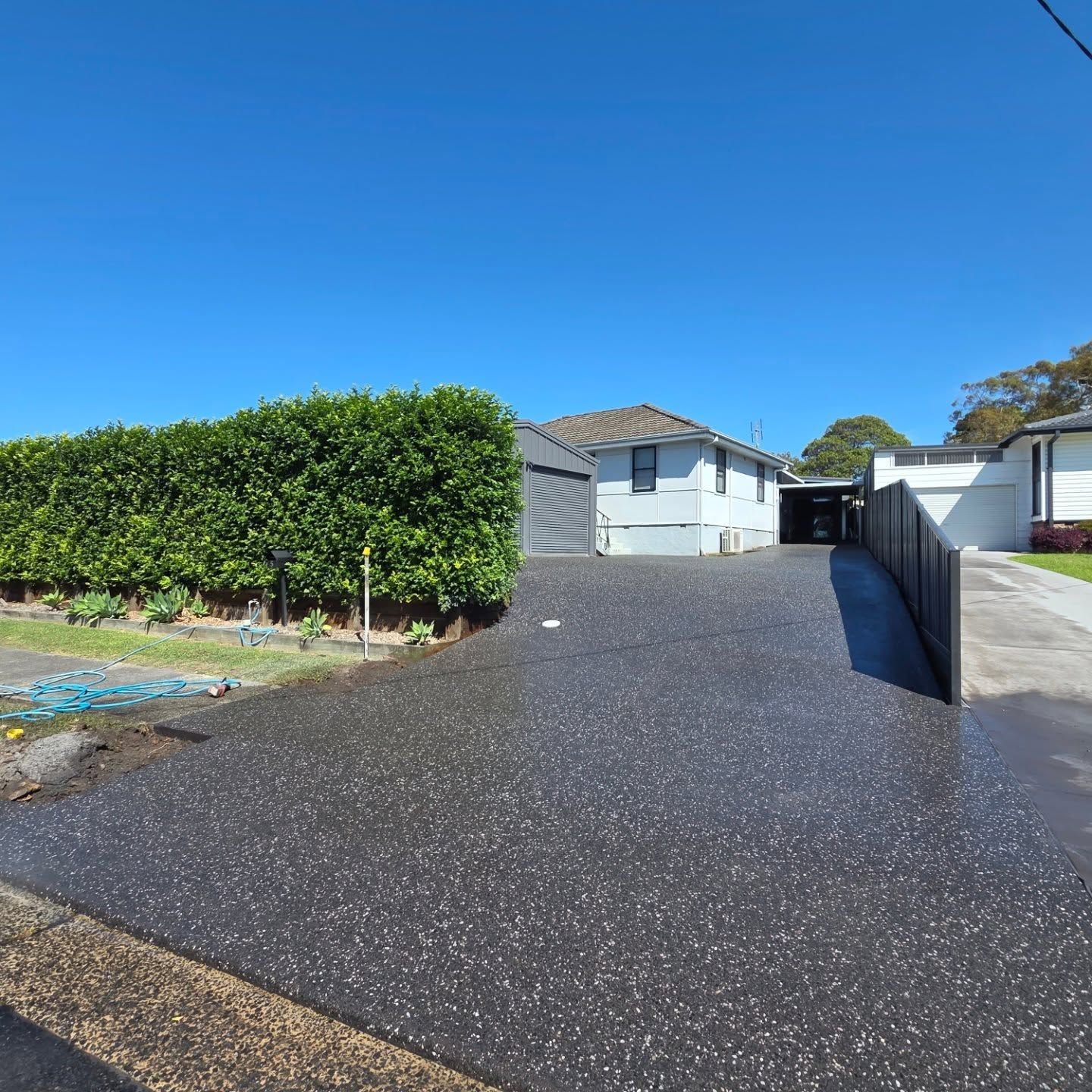 A dark driveway with light-colored aggregate leads toward a white house next to a tall, manicured green hedge. — JDB Concreting in Wollongong, NSW