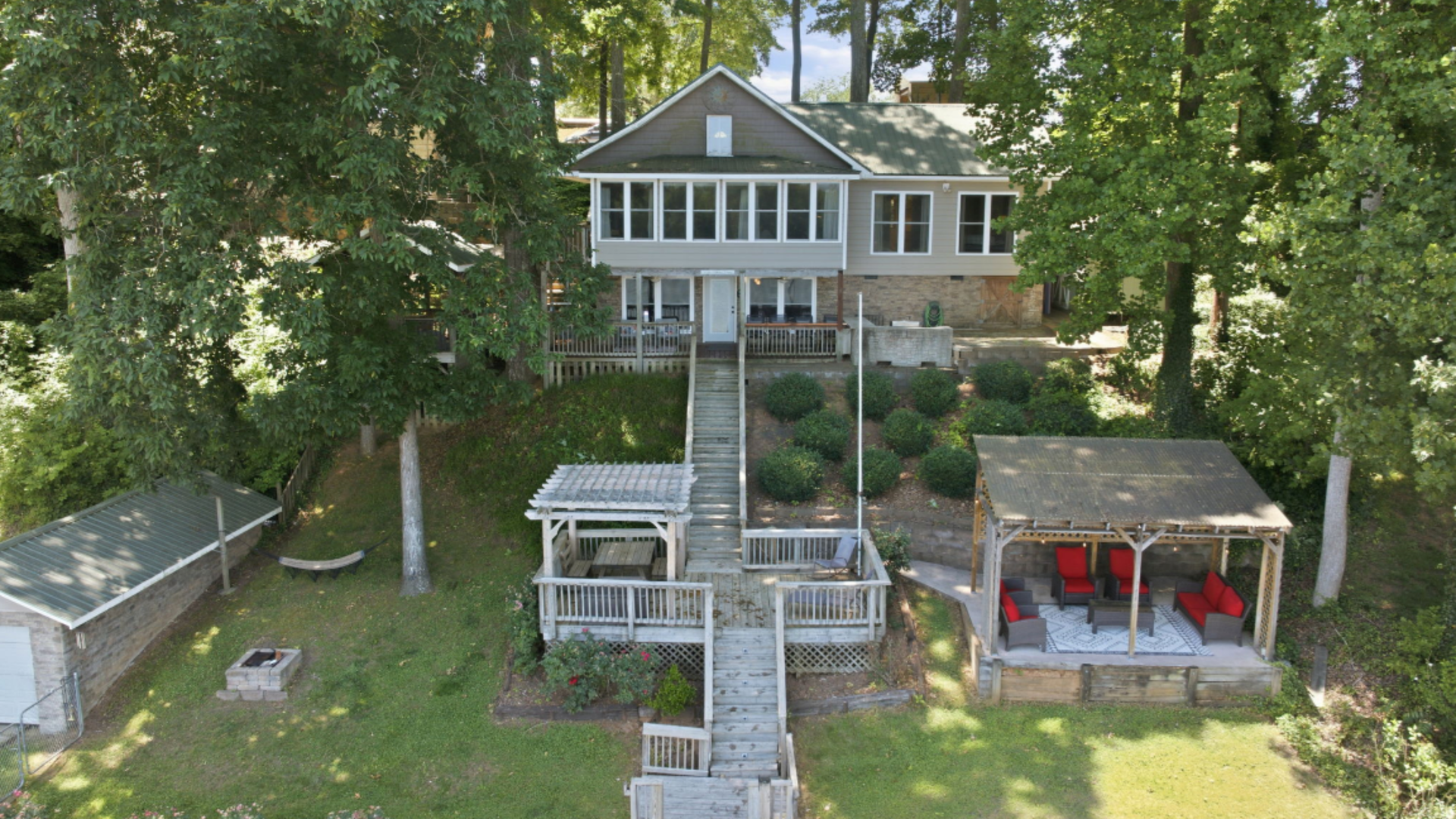 Aerial view of a lakeside house with a long wooden dock, trees, and two covered seating areas.