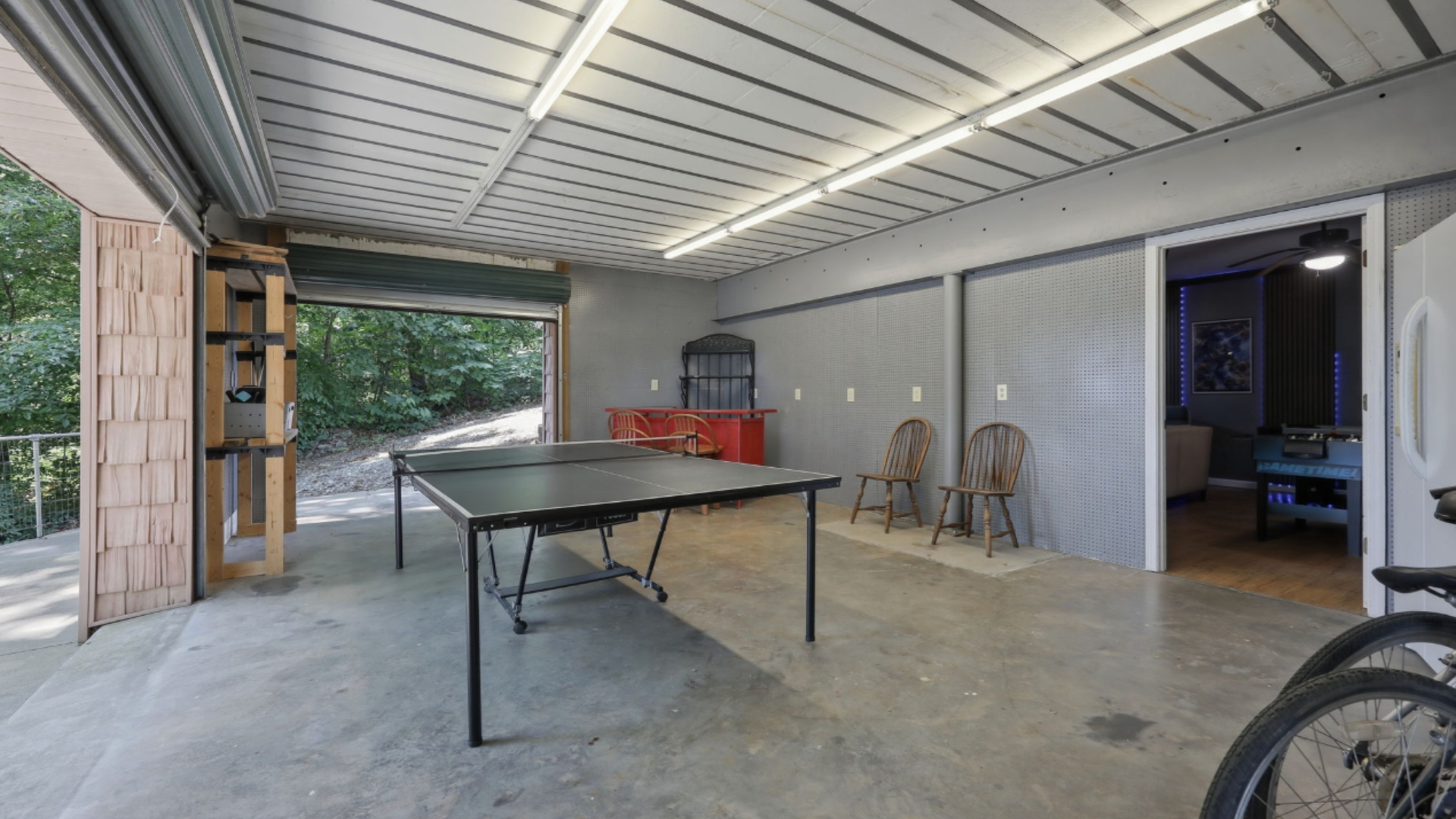 Covered patio with a ping-pong table, folding chairs, and a red cooler beside open doors to a garage.