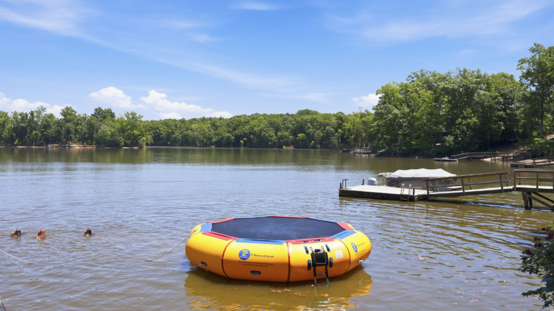Yellow inflatable water trampoline on a calm lake with swimmers, dock, and trees under a blue sky