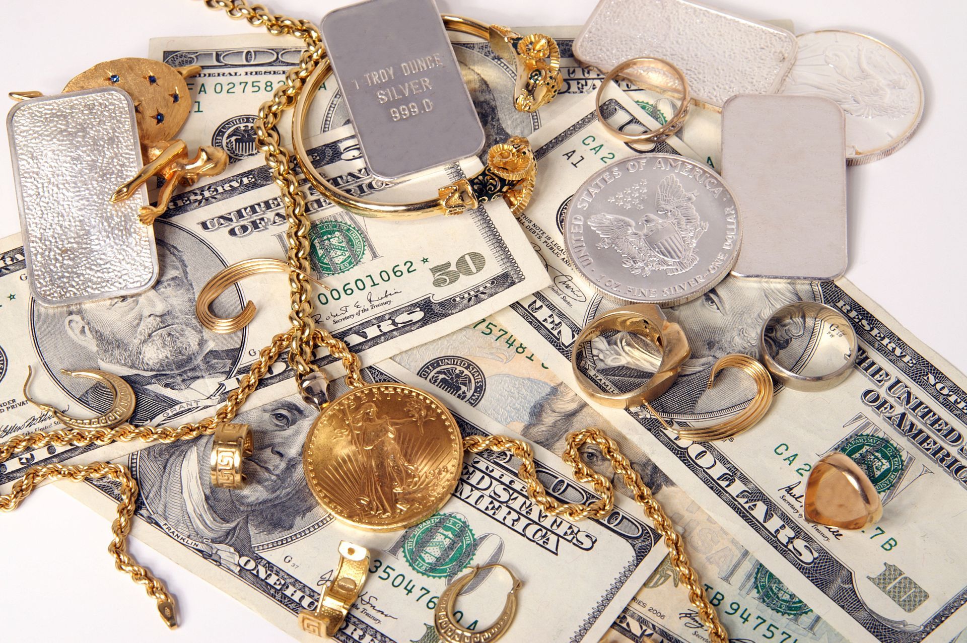 Gold jewelry, silver coins, and money (US $100 bills) scattered on a white surface.