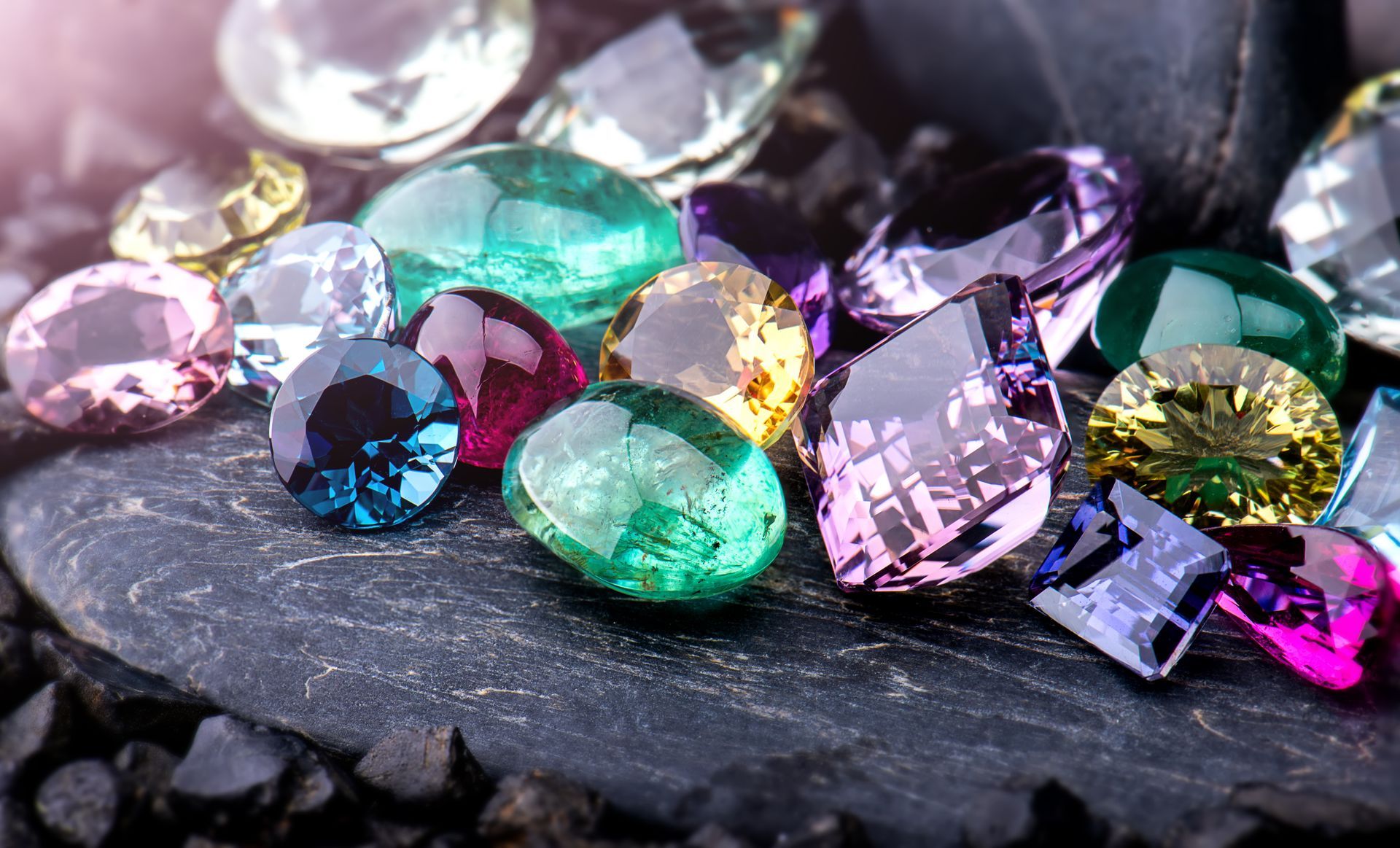 Colorful assortment of faceted gemstones including diamonds, sapphires, and emeralds on dark slate