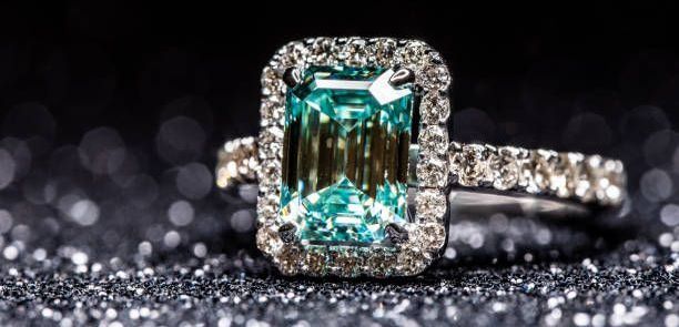 Emerald-cut ring with a turquoise gemstone and diamond halo, set on a sparkly, dark background.