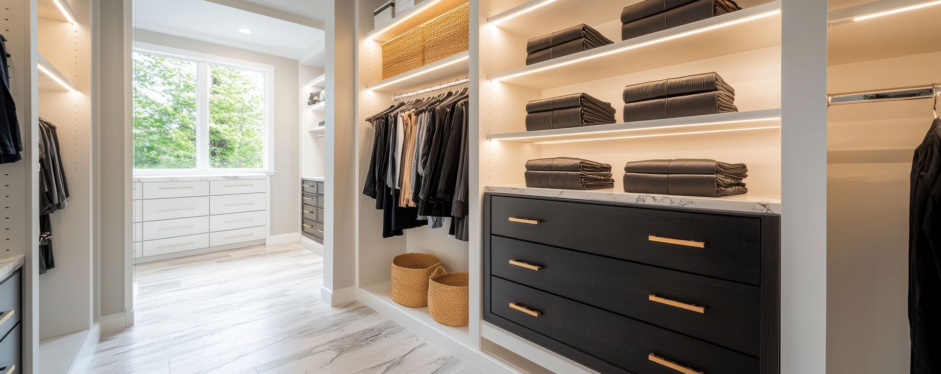 Un dressing moderne et lumineux, avec des placards blancs, des tiroirs en bois foncé, des vêtements suspendus et une fenêtre lumineuse.