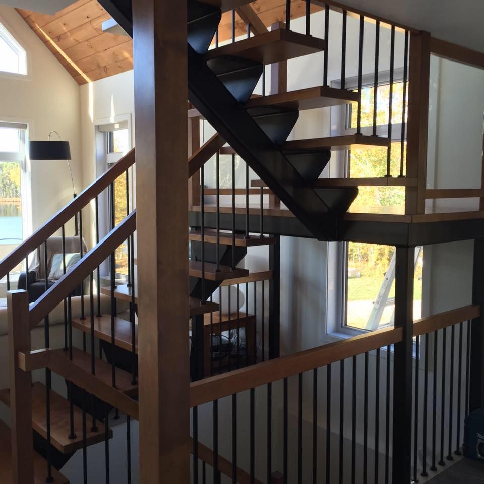 Un escalier intérieur moderne avec marches en bois, rampes et supports en métal noir, dans une pièce baignée de lumière naturelle avec plafond en bois.