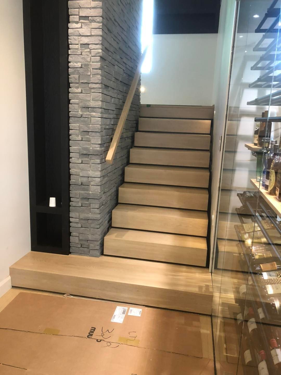Escalier en bois avec un mur en pierre apparente et un casier à vin vitré sur la droite, sol intérieur recouvert de carton.
