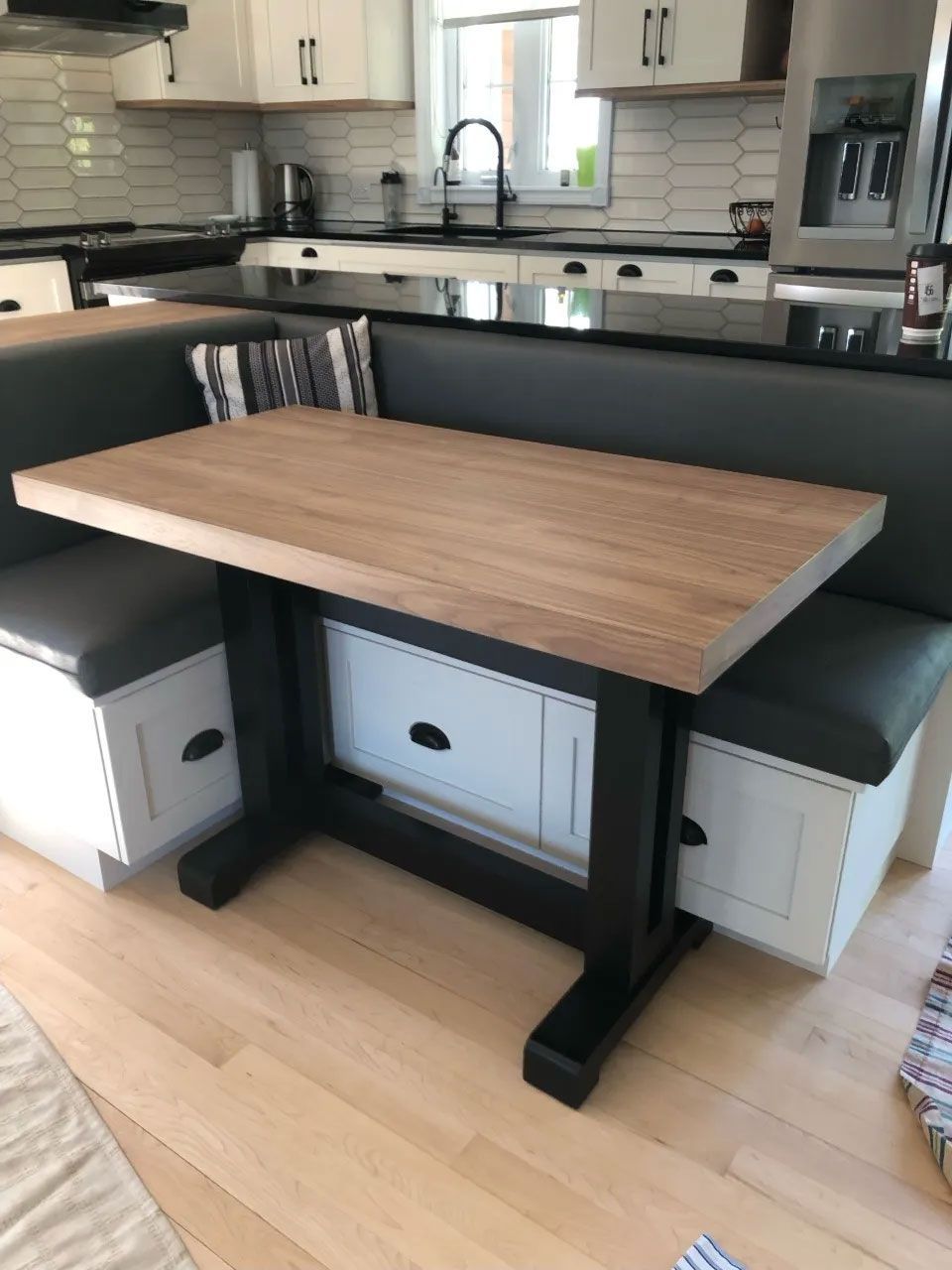 Un coin cuisine avec une table à plateau en bois et un pied central noir, placée entre une banquette grise rembourrée avec tiroirs.