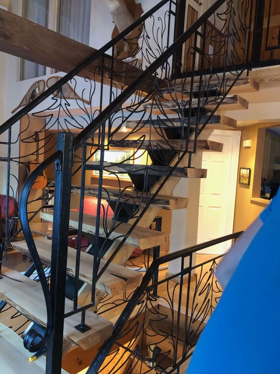 Un escalier avec des marches en bois et une rampe décorative en fer noir ornée d'un motif en forme d'arbre, à l'intérieur d'une maison.