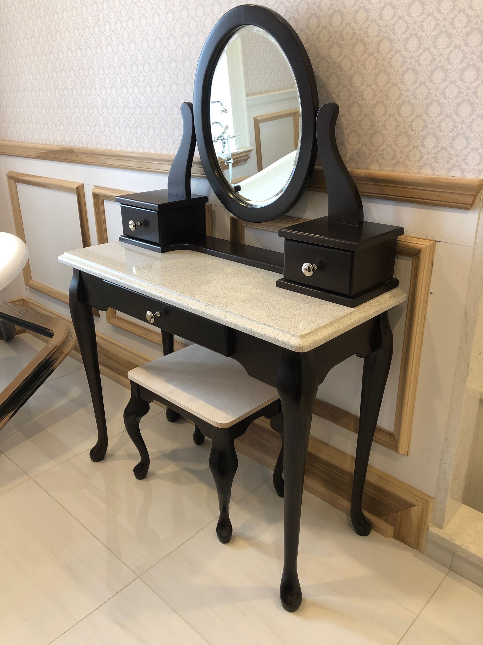 Une coiffeuse noire avec un plateau en marbre, un miroir ovale, deux petits tiroirs et un tabouret rembourré assorti, adossée au mur.