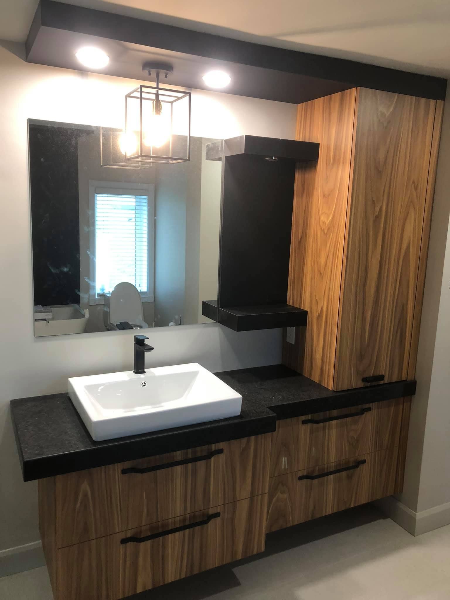 Meuble-lavabo moderne avec armoires en bois, comptoir noir, vasque blanche et luminaire à cadre noir.