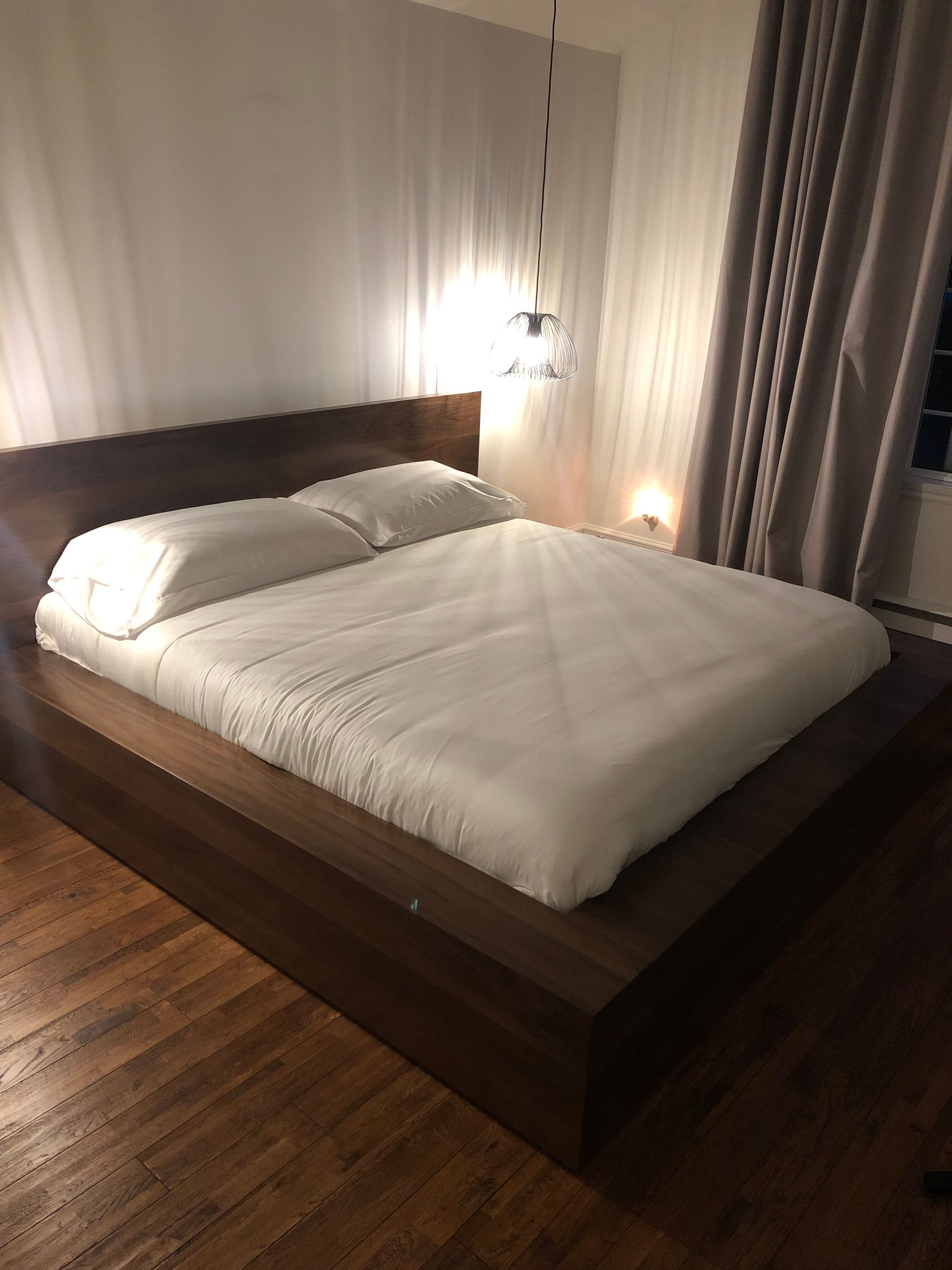 Un lit plateforme en bois bas avec du linge de lit blanc, placé dans une chambre avec parquet et rideaux.