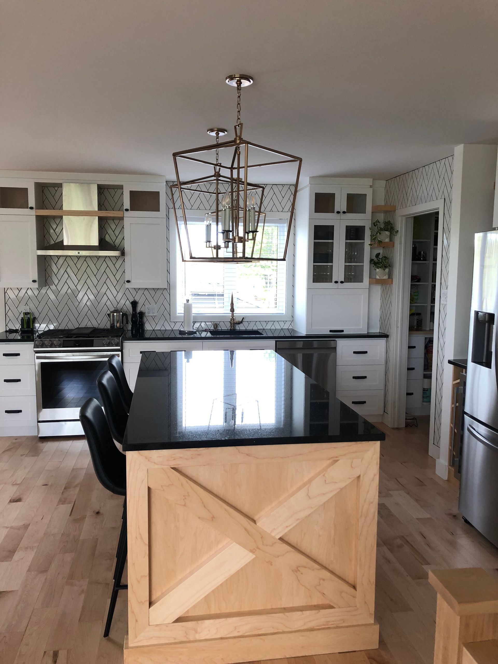 Une cuisine avec un îlot en bois naturel, des plans de travail noirs, des placards blancs, un parquet clair et un luminaire en forme de lanterne métallique.