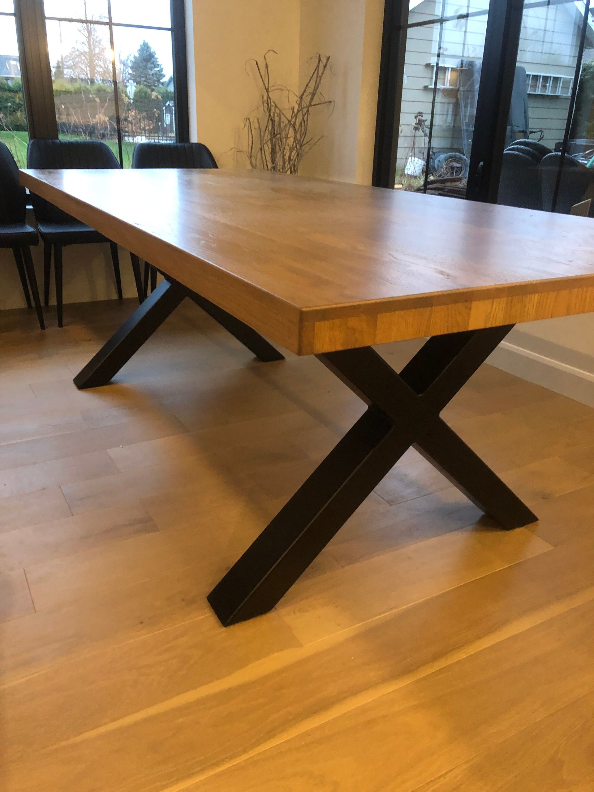 Une table à manger avec un plateau épais en bois clair et des pieds en métal noir en forme de X, dans une salle à manger moderne.