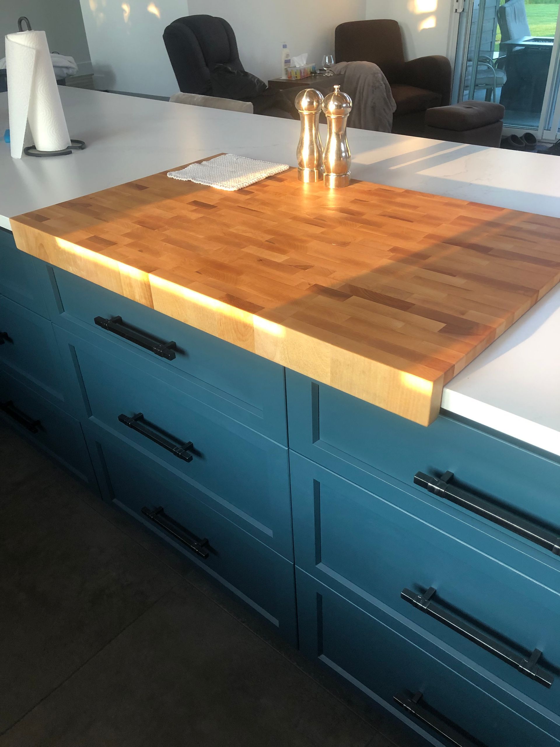Une grande planche à découper en bois massif trône sur un îlot de cuisine bleu turquoise aux poignées noires ; un rouleau d'essuie-tout se trouve à proximité.