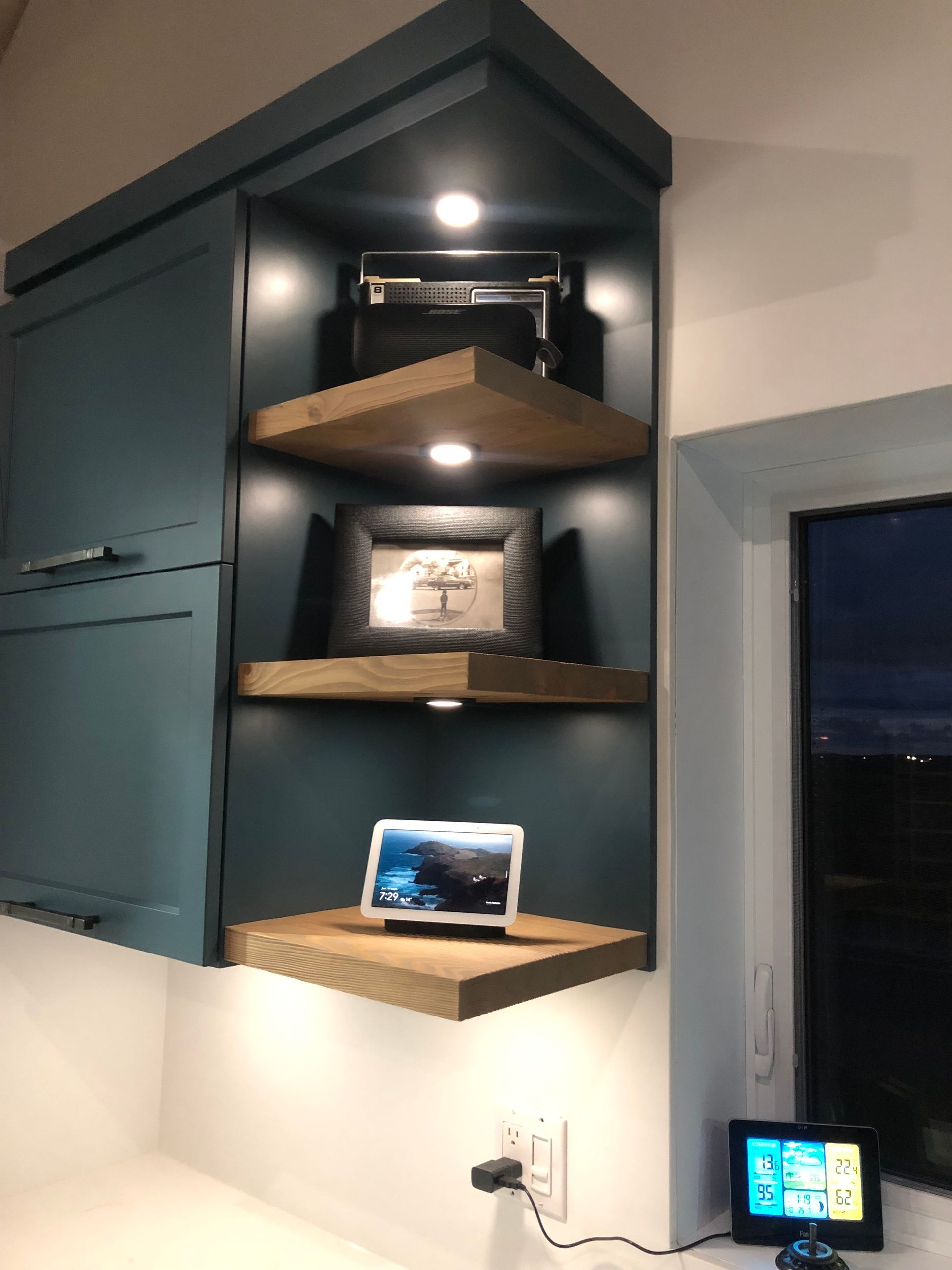 Meuble d'angle bleu sarcelle foncé avec trois étagères en bois éclairées présentant un appareil photo vintage, une photo encadrée et un écran intelligent.