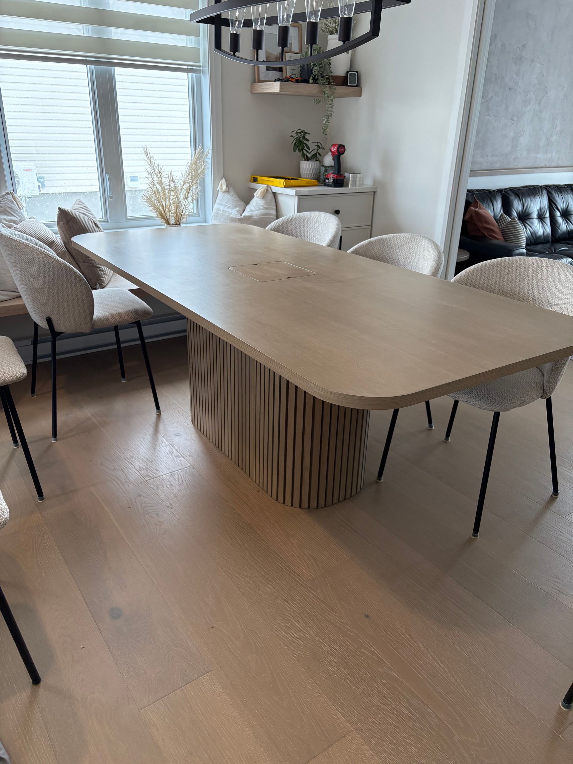 Une table de salle à manger en chêne clair avec un piètement texturé et cannelé, entourée de chaises modernes rembourrées sur un parquet.