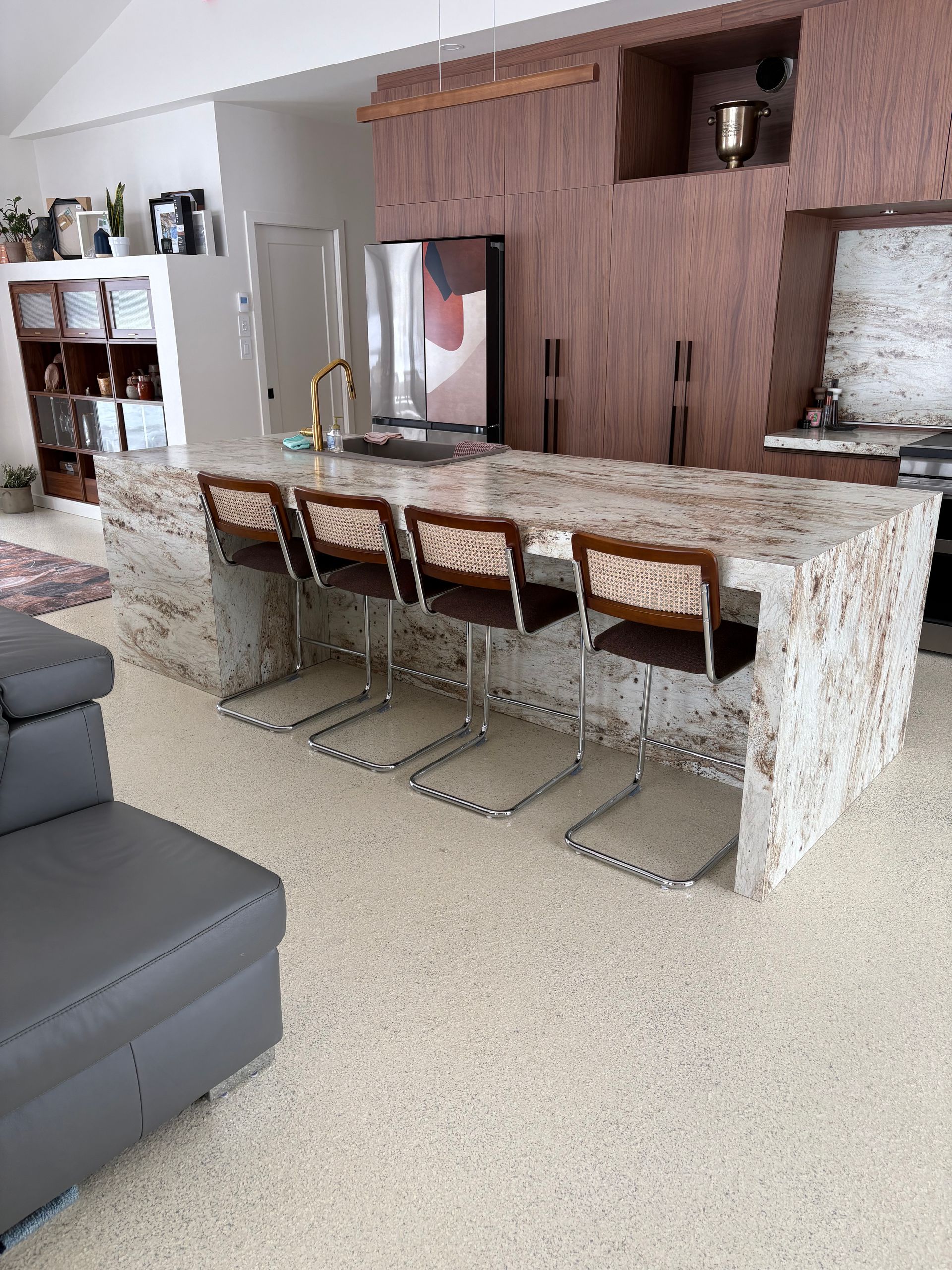 Un grand îlot de cuisine en pierre avec quatre tabourets modernes, placé devant des armoires en bois foncé et des appareils électroménagers en acier inoxydable.