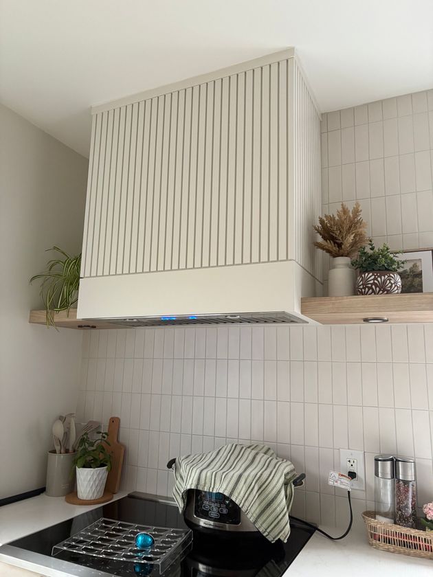 Une hotte aspirante verticale à lamelles couleur crème au-dessus d'une plaque de cuisson, avec des murs carrelés de blanc et des étagères flottantes en bois.