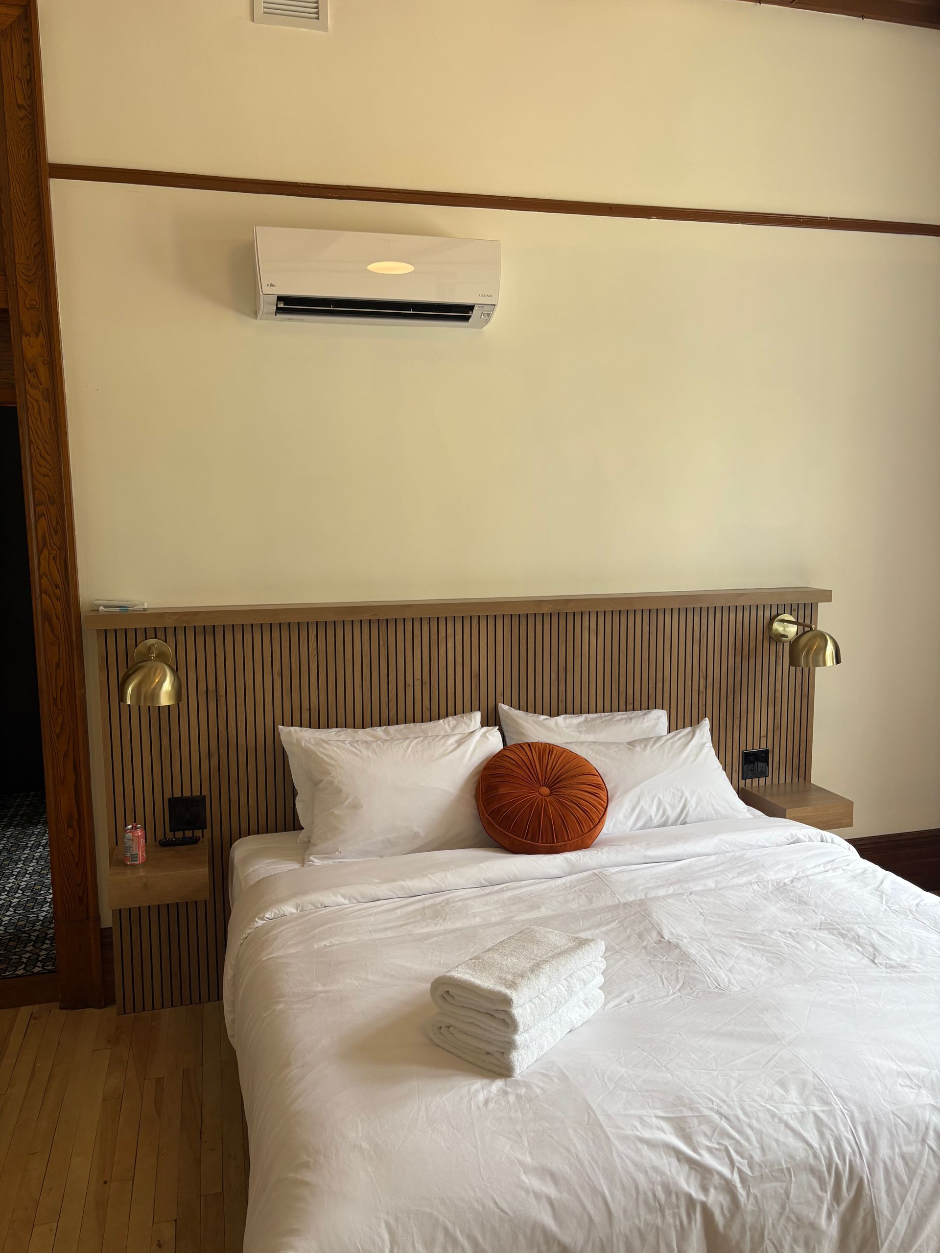 Un lit blanc avec des serviettes pliées et un oreiller rond orange, adossé à une tête de lit en bois à lattes, sous un climatiseur.