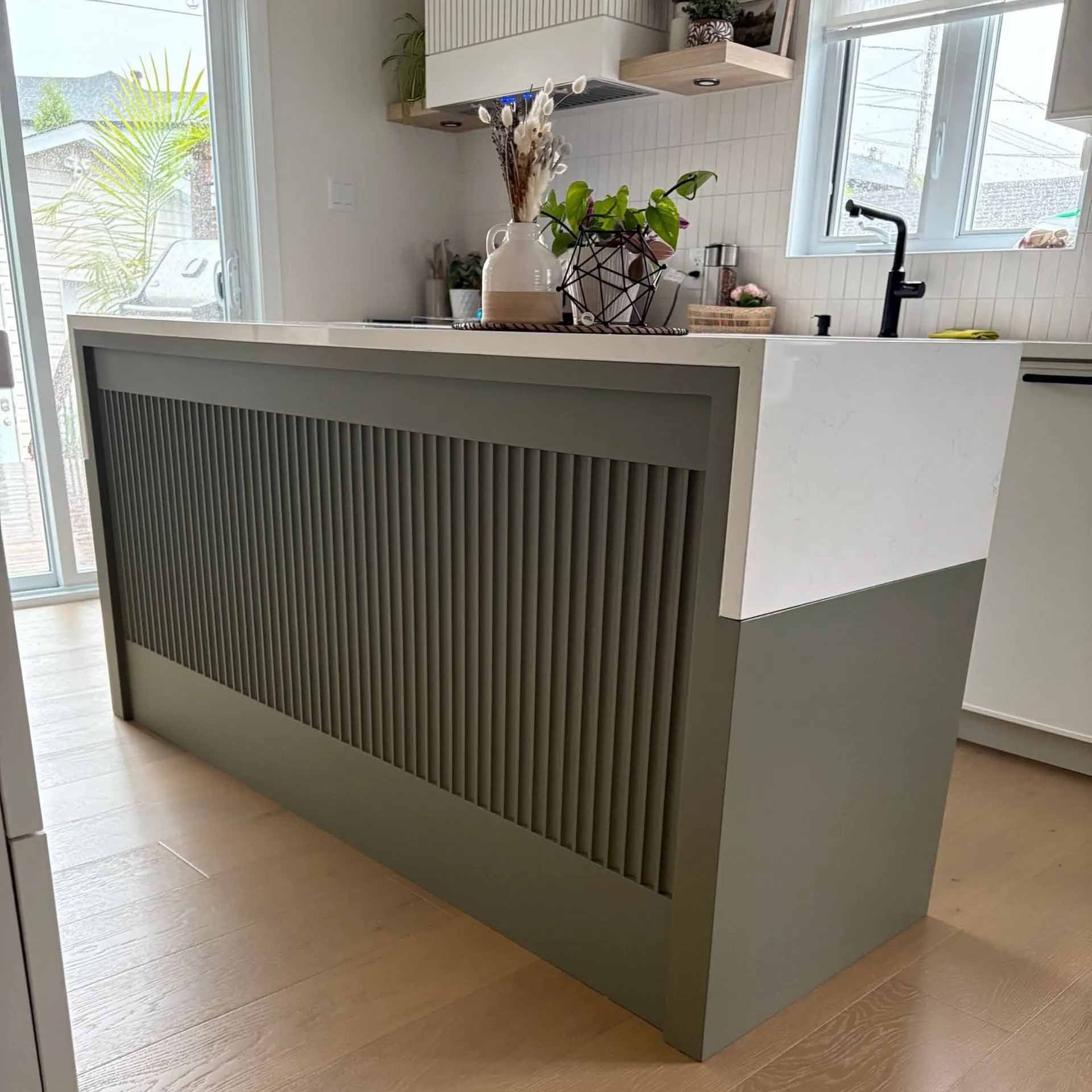 Un îlot de cuisine avec une base gris foncé ornée de lattes verticales, surmontée d'un plan de travail blanc et de petits objets décoratifs.