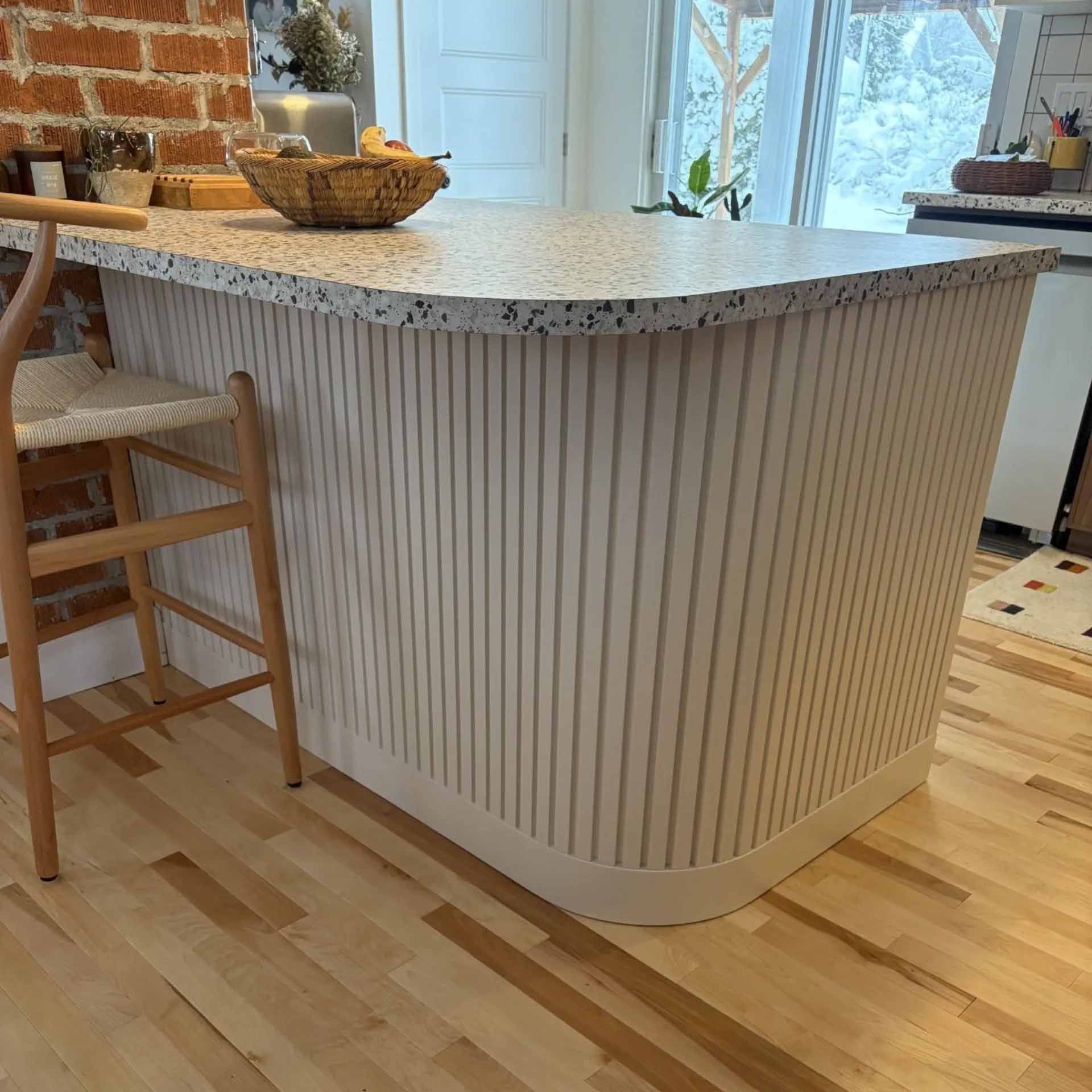 Un îlot de cuisine clair avec une base incurvée et cannelée et un plan de travail moucheté, à côté d'un tabouret de bar en bois.