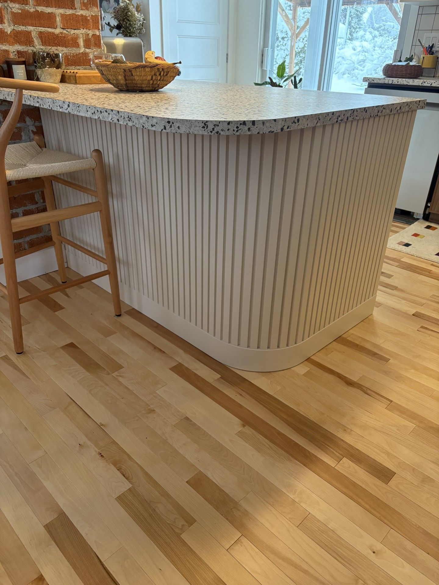 Un îlot de cuisine avec une base incurvée à lattes verticales blanches et beiges et un plan de travail moucheté, sur un parquet clair.