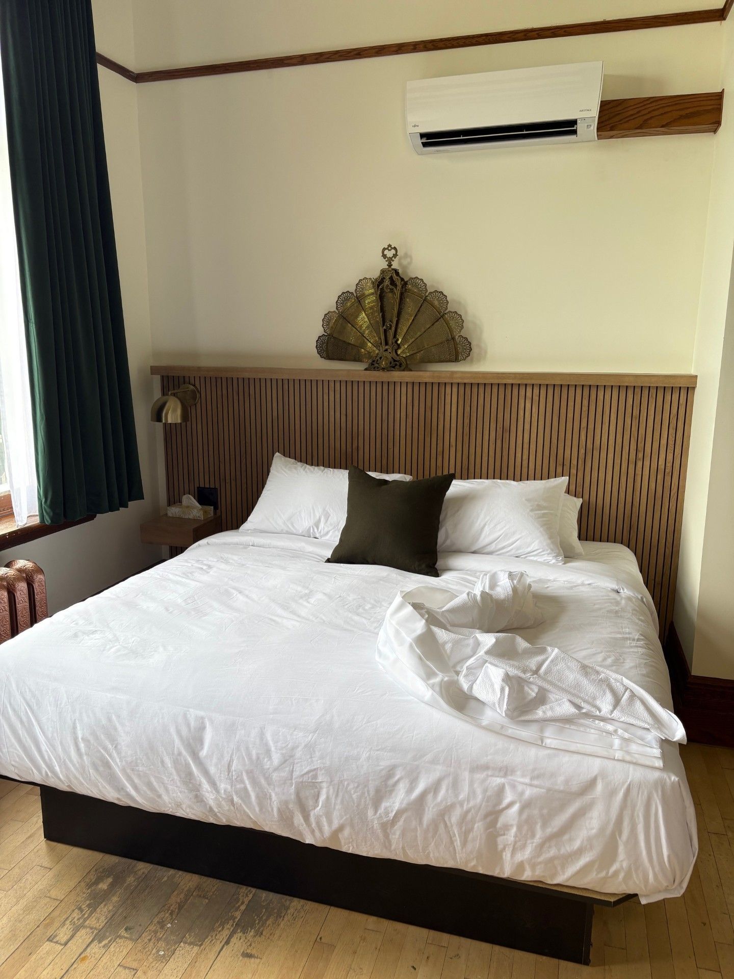 Un lit queen size avec une couette blanche, une tête de lit en bois, une décoration murale représentant un éventail doré et un climatiseur fixé sur un mur blanc.