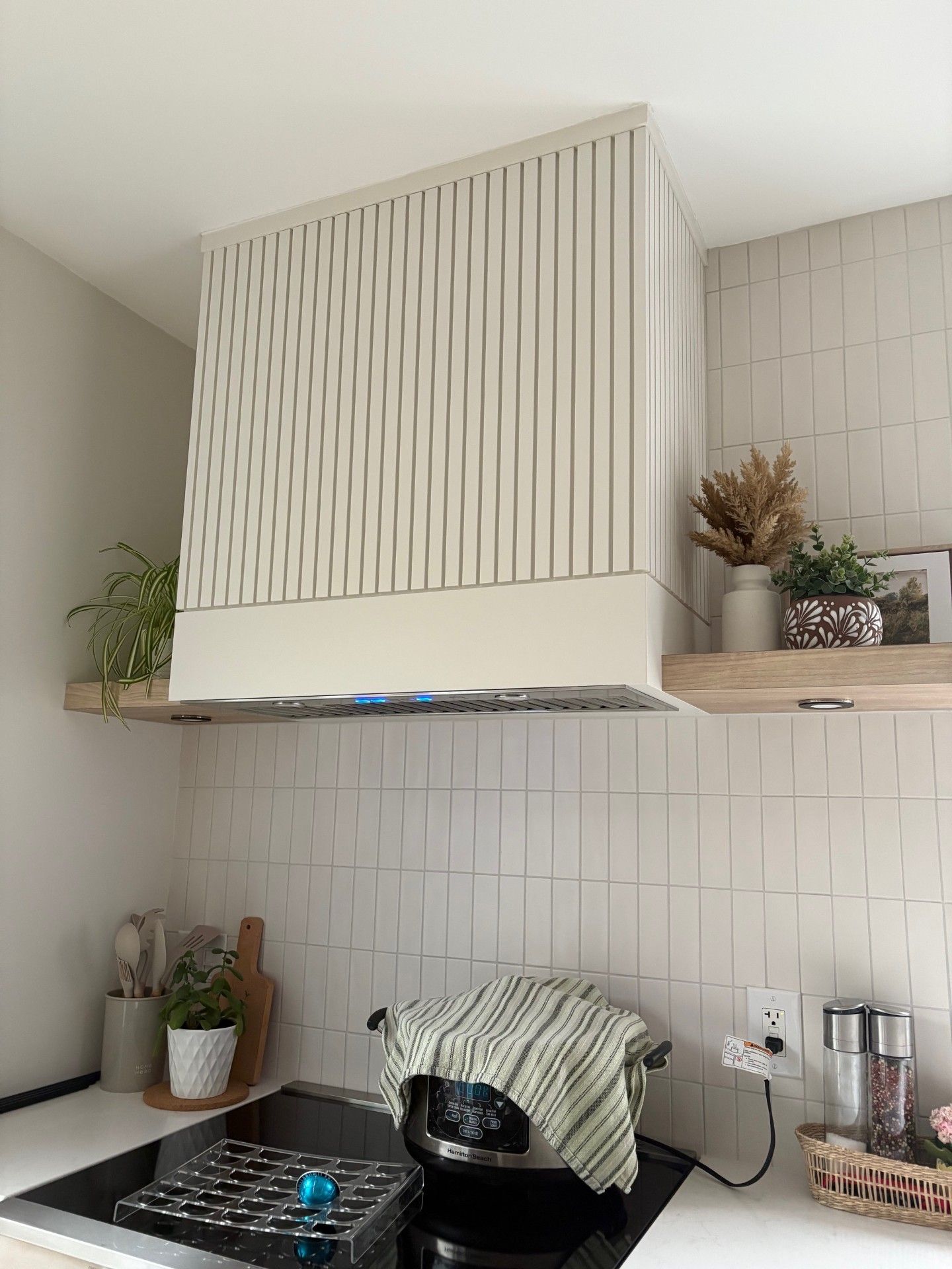 Un coin cuisine avec une hotte aspirante haute, claire et à lamelles verticales au-dessus d'une cuisinière et un mur carrelé de blanc.