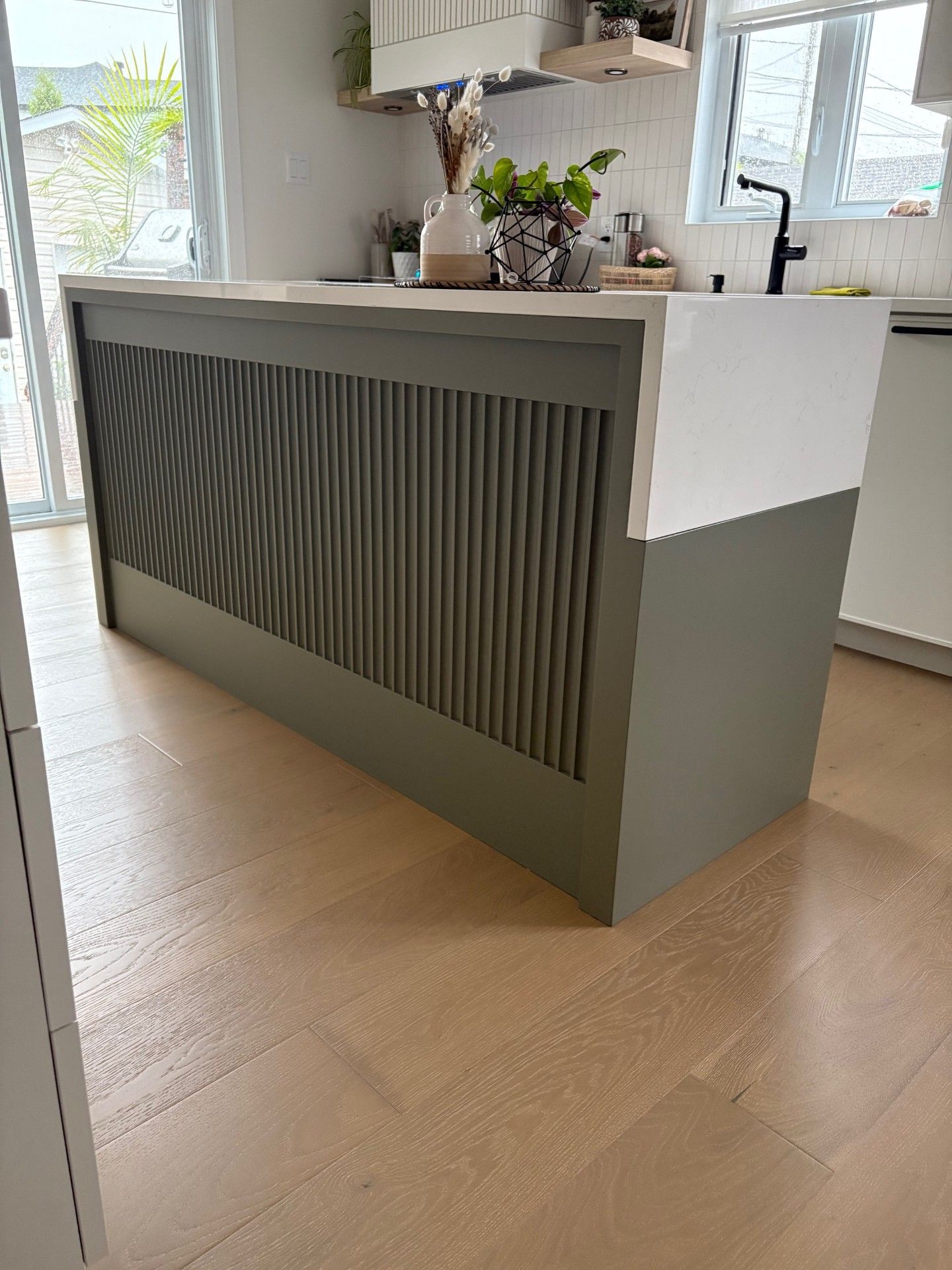 Un îlot de cuisine avec un panneau avant à lattes verticales vert sauge foncé et un plan de travail blanc, installé sur un parquet clair.