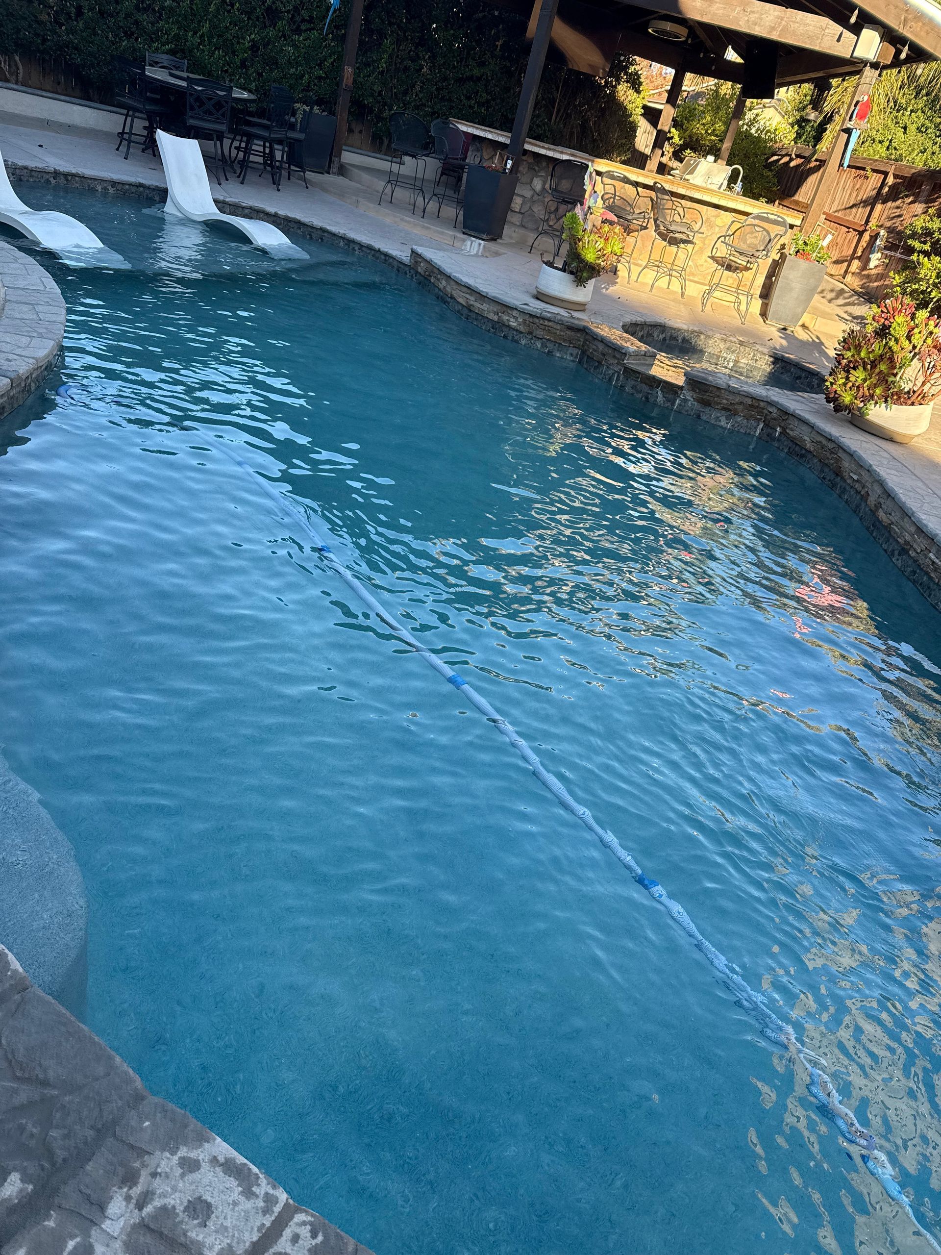 Swimming pool with blue water and stone surround, surrounded by outdoor furniture and plants.