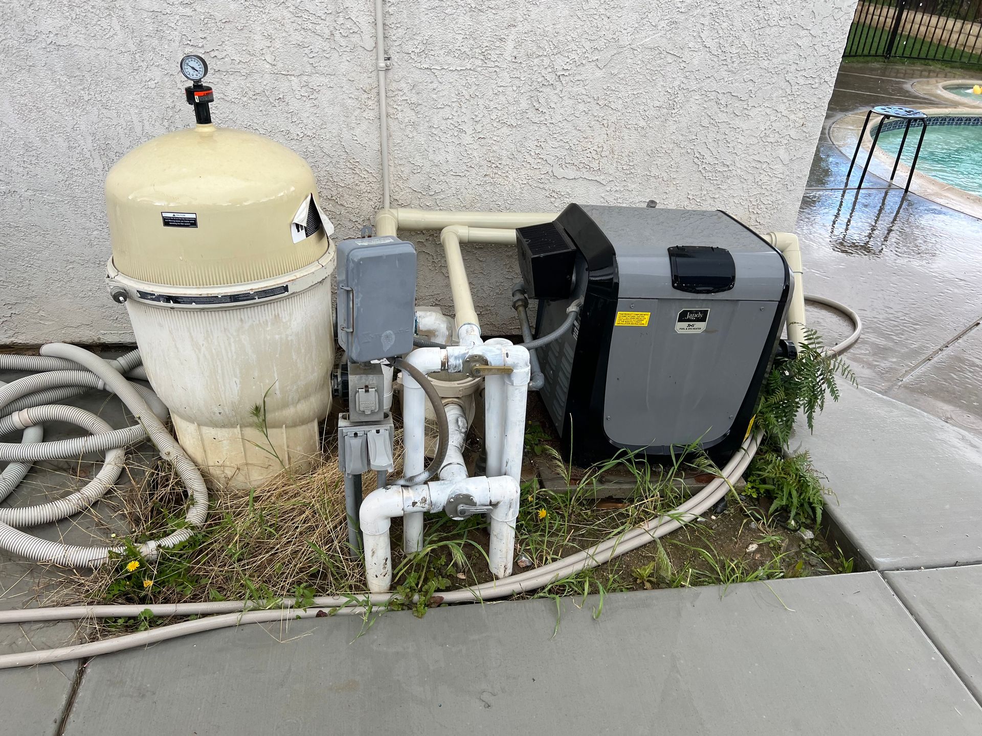 Pool equipment with filter, pipes, and pump beside a wet concrete walkway outdoors