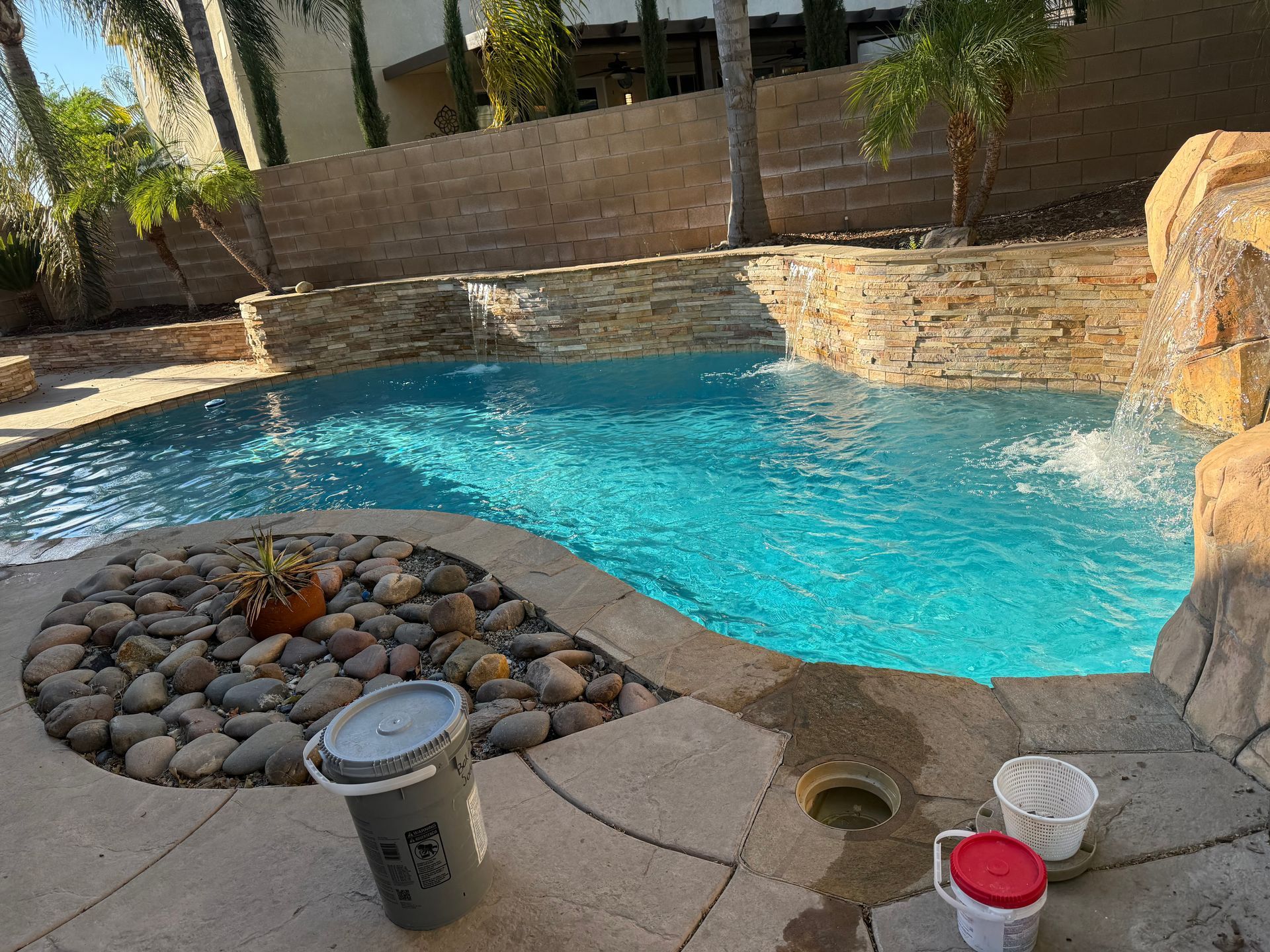 Swimming pool with a waterfall and stone landscaping, with buckets visible nearby.