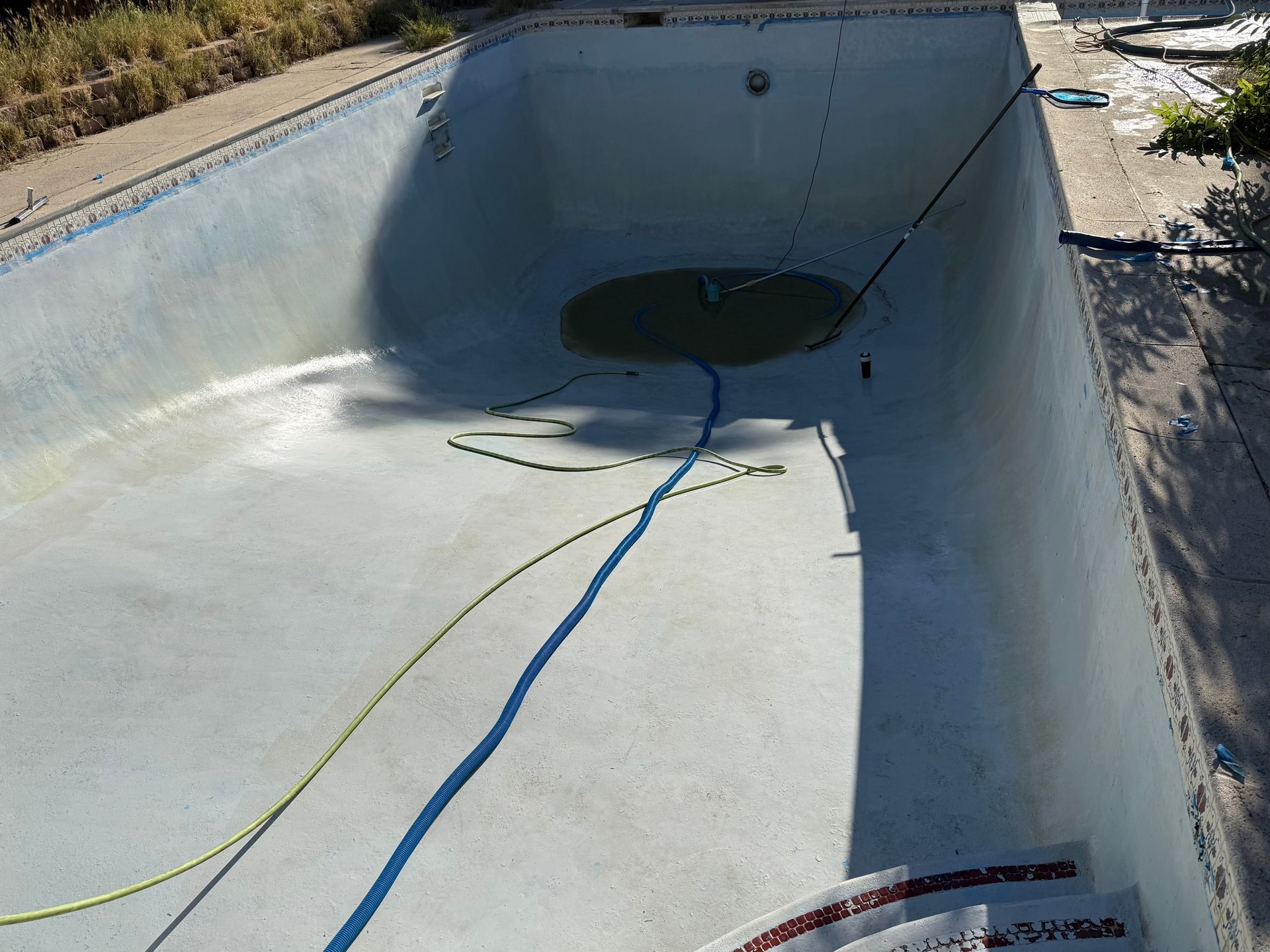 Empty concrete pool with a circular drain, hoses on the floor, and bright sunlight casting shadows