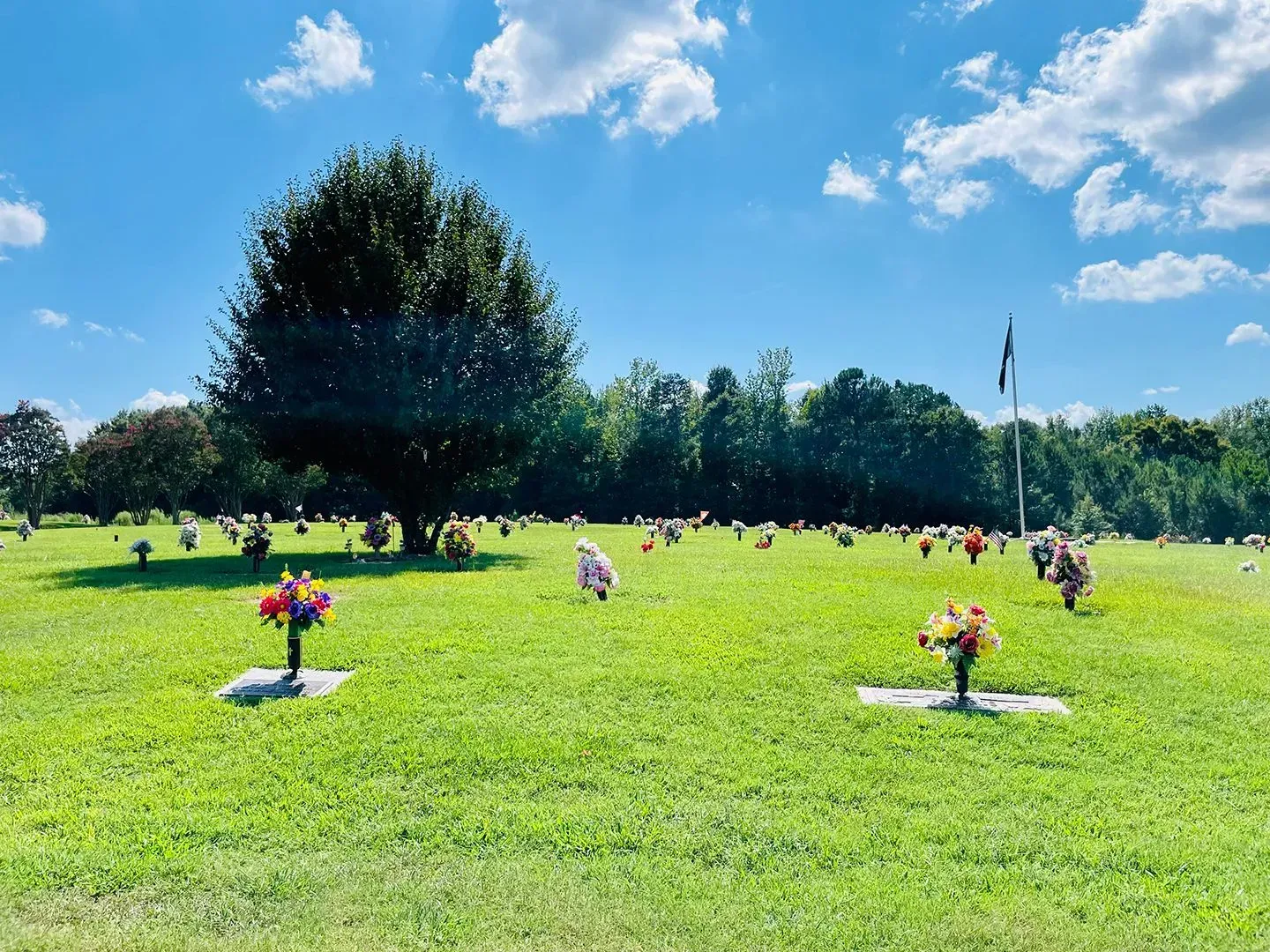 Wide green cemetery lawn with flower arrangements and white grave markers under a bright blue sky