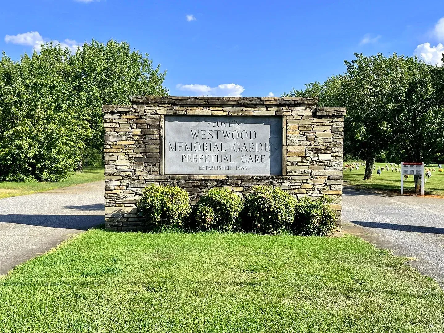 Stone entrance sign for Westwood Memorial Gardens, surrounded by green grass and trees under a blue sky.