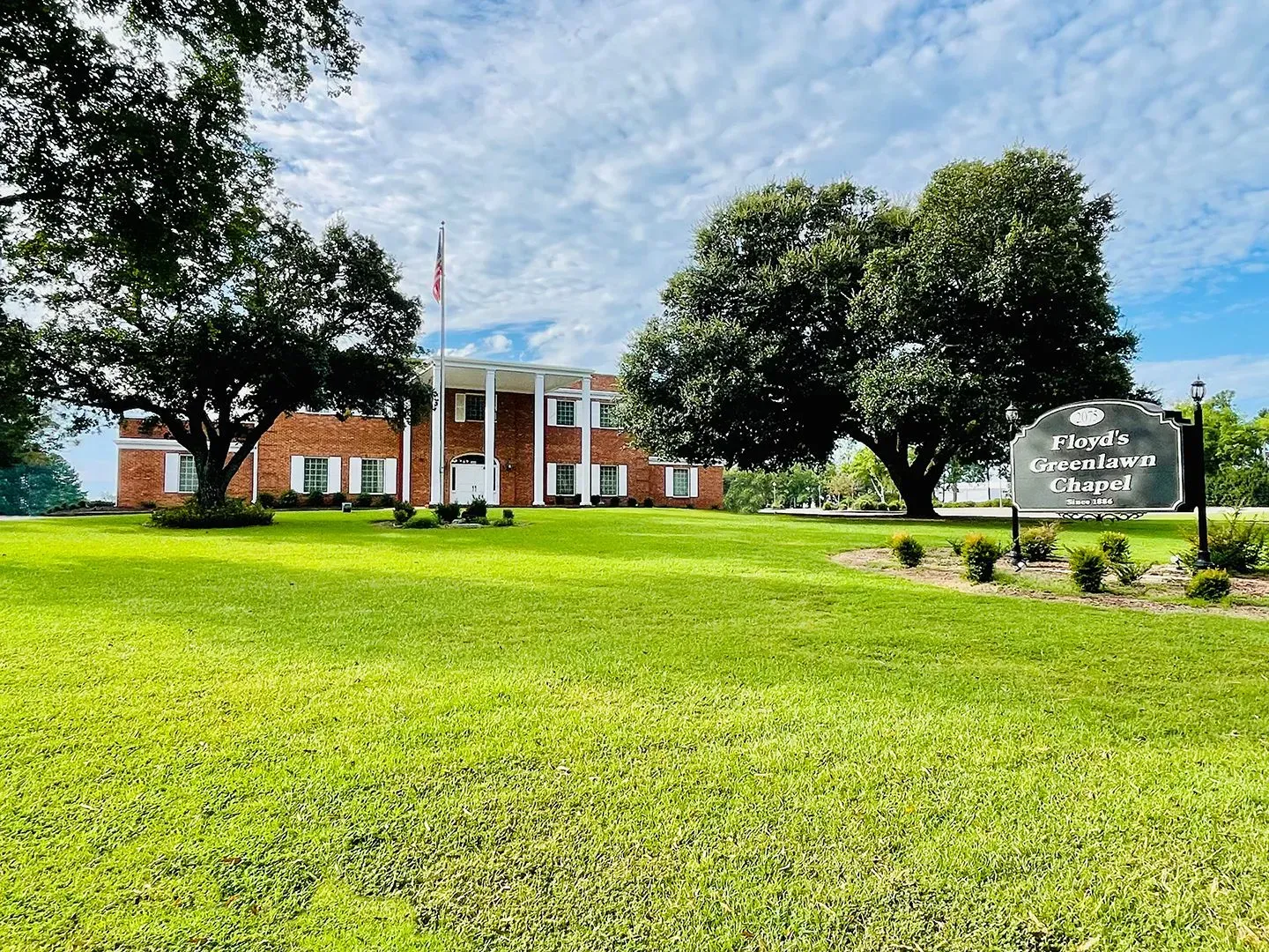 Lawns and trees in front of a red-brick building with white columns and a campus sign.
